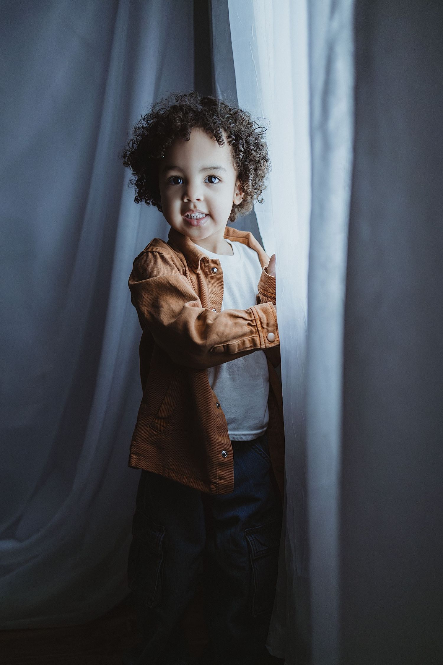 Adorable curly-haired toddler in brown jacket peeks through white curtains with a playful smile.