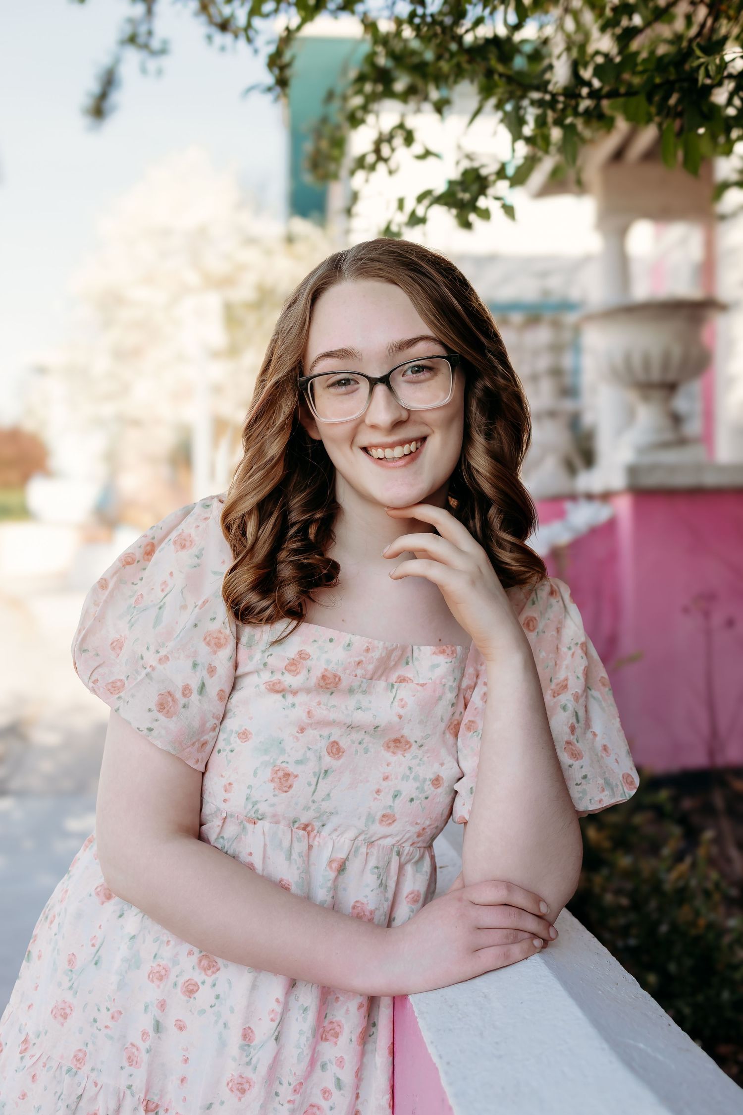 Smiling teenage girl with glasses in floral pink dress leaning on white railing outdoors.