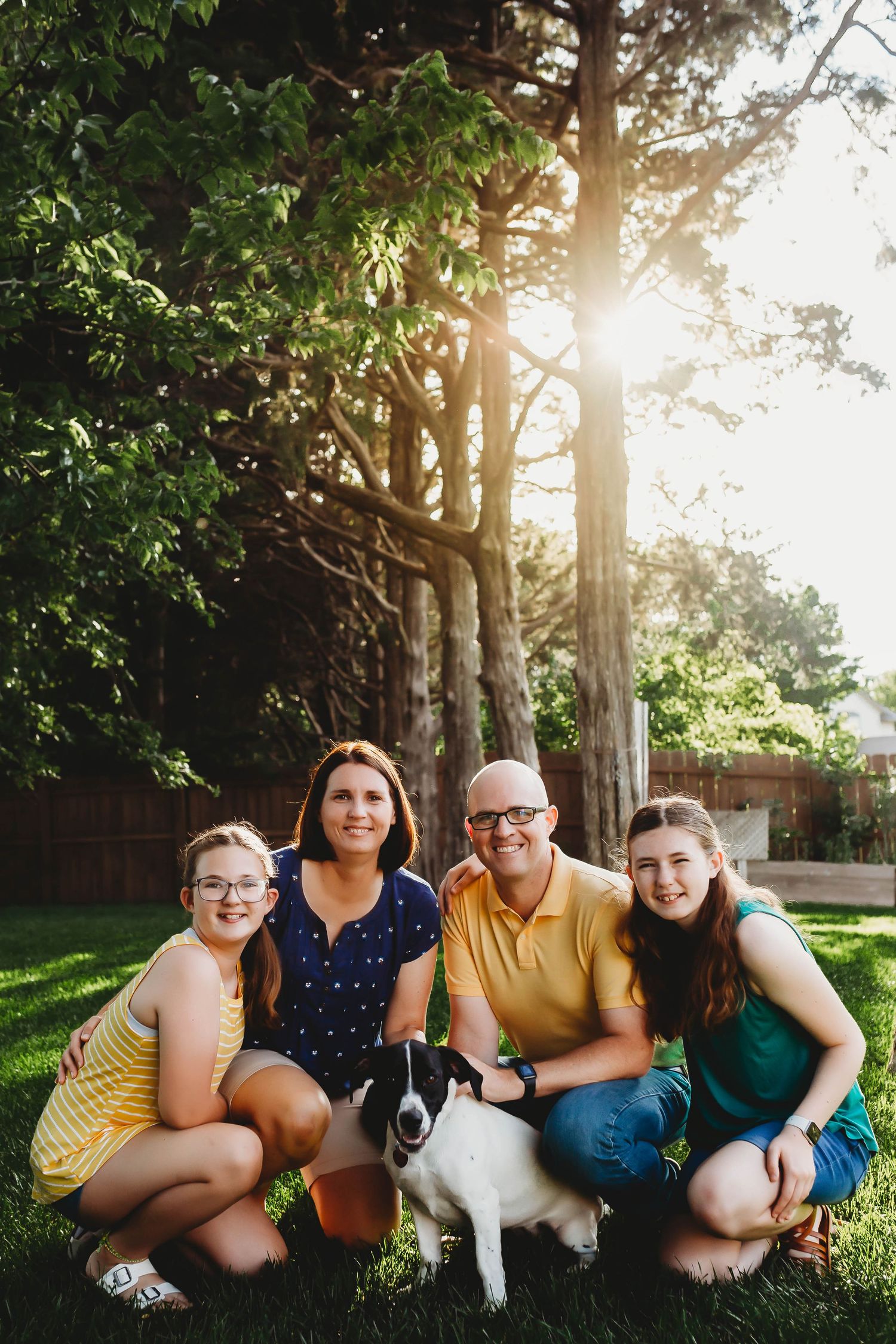A happy family of four poses with their black and white dog in a sunlit backyard surrounded by tall trees.