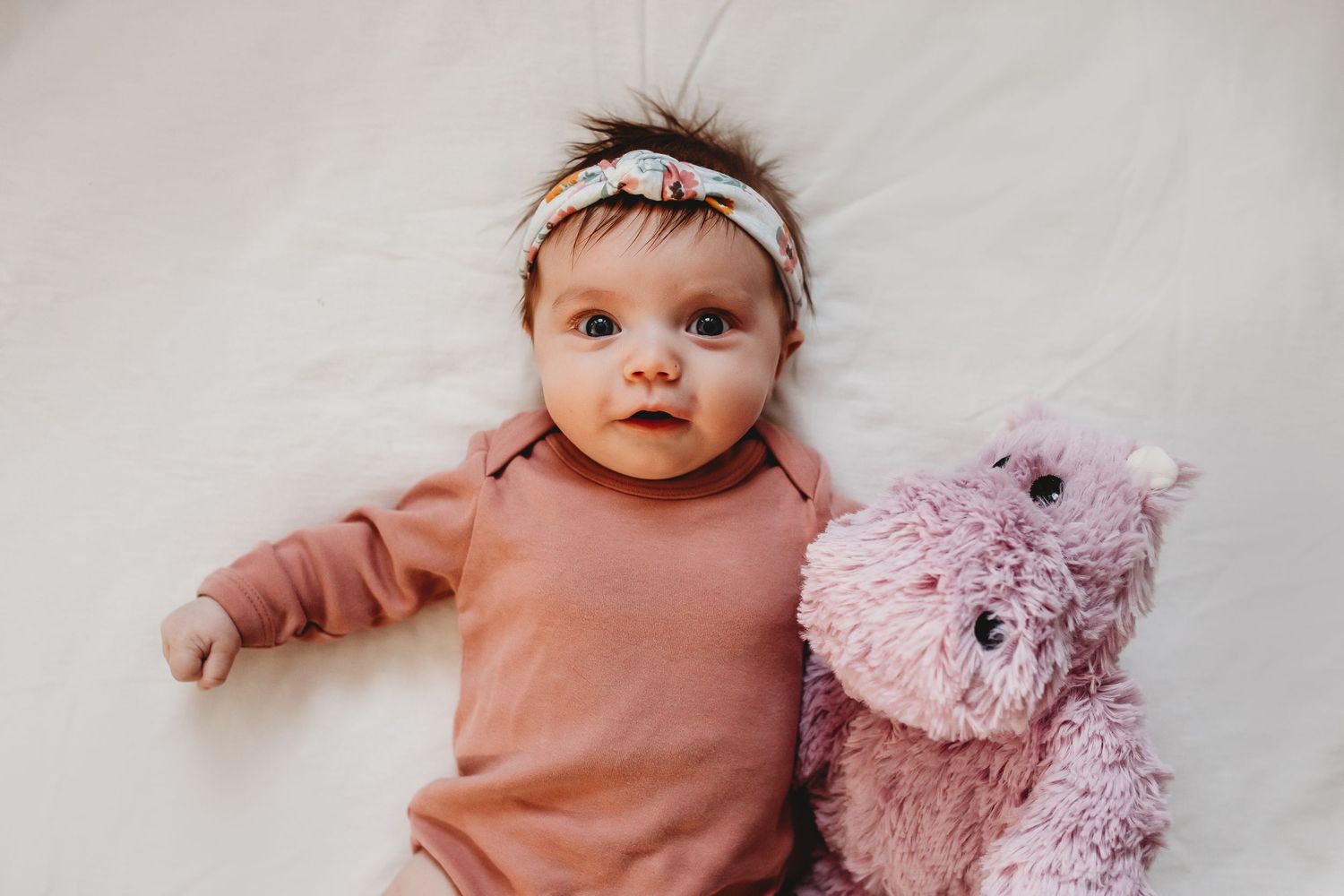 Adorable baby girl in pink outfit lying next to a fluffy pink teddy bear with a floral headband.