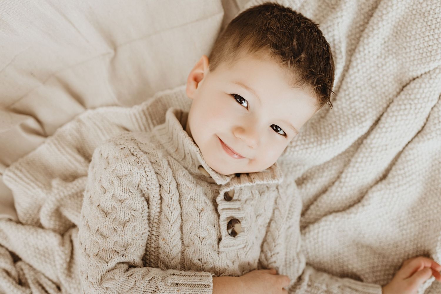 Smiling toddler boy in cream cable-knit sweater lying on a cozy beige blanket.
