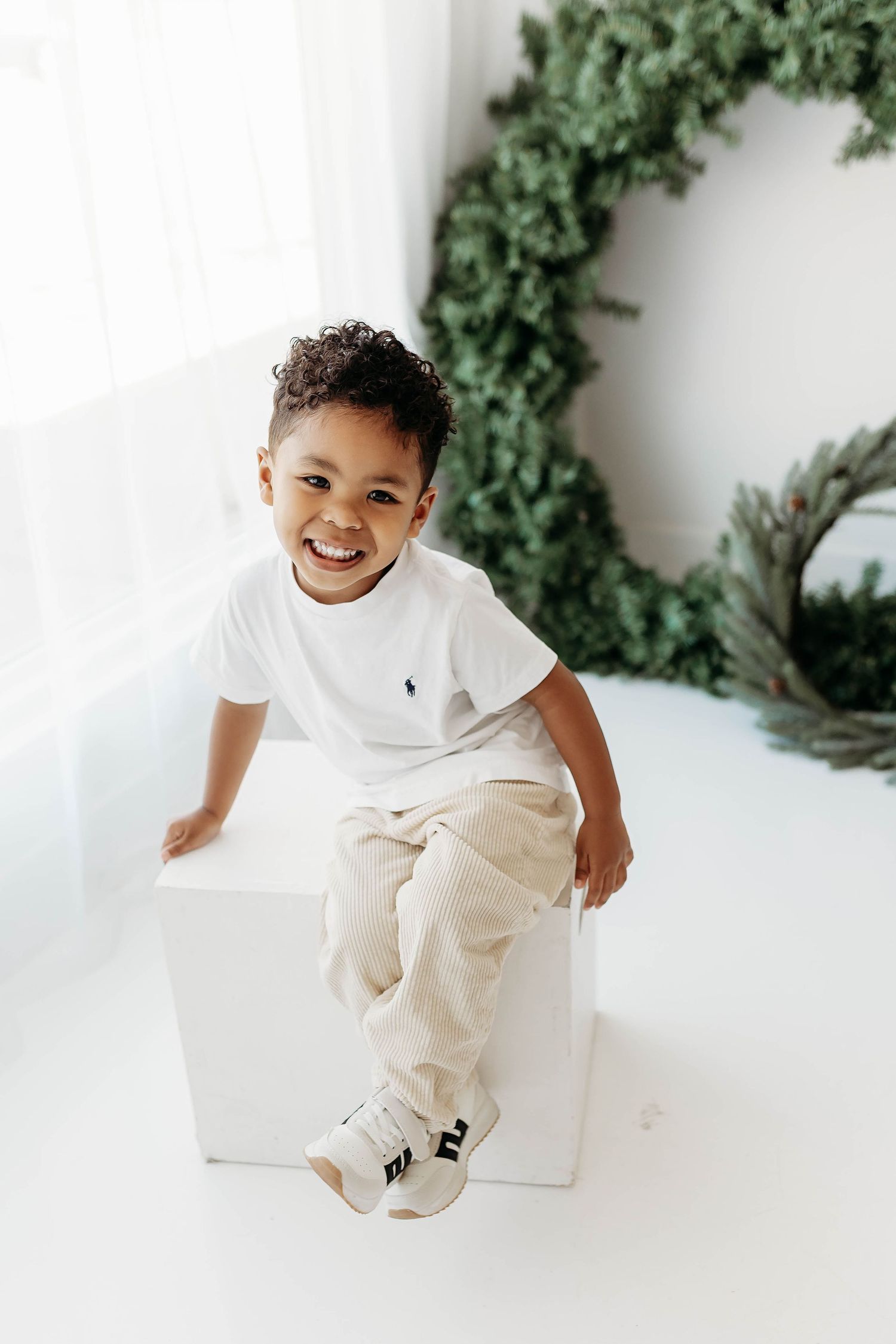 A happy young boy in a white polo shirt sits on a white cube smiling in a bright studio.