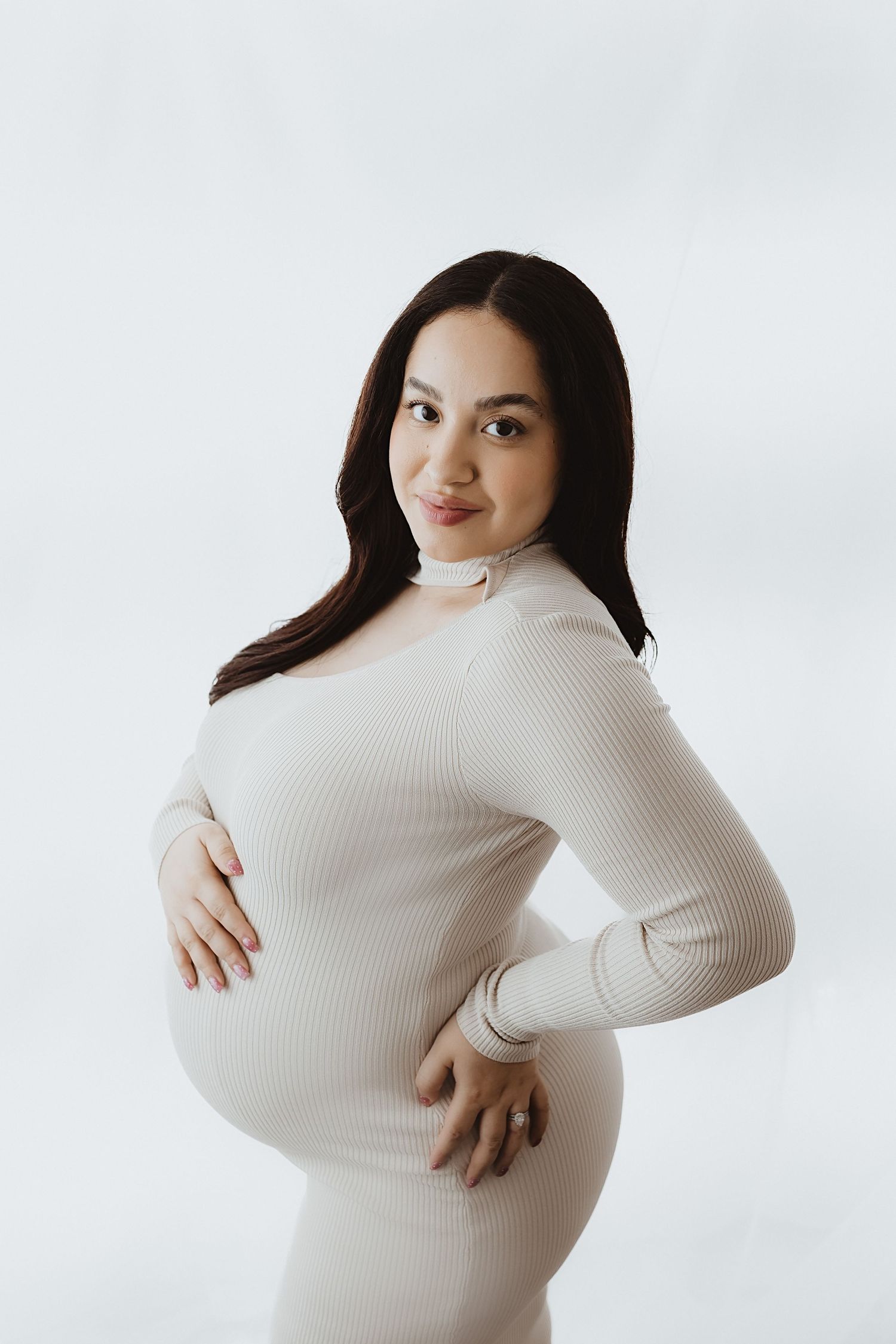 Pregnant woman in white ribbed dress cradling baby bump, smiling softly against light background.