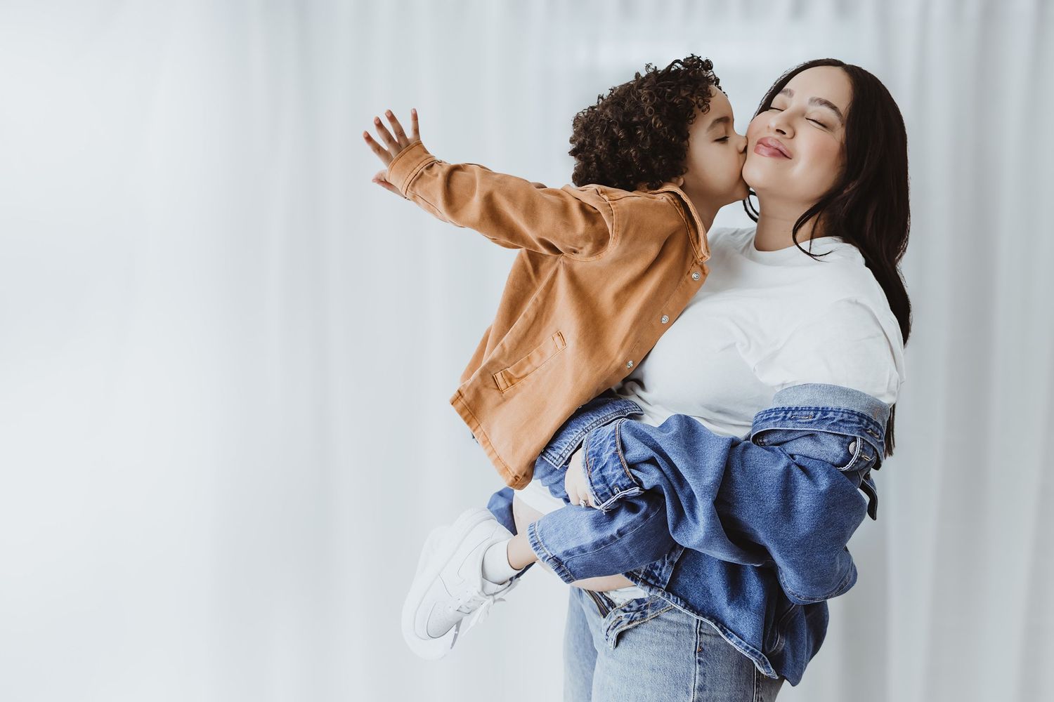 A mother holds her young child who gives her a kiss on the cheek while spreading arms wide indoors.