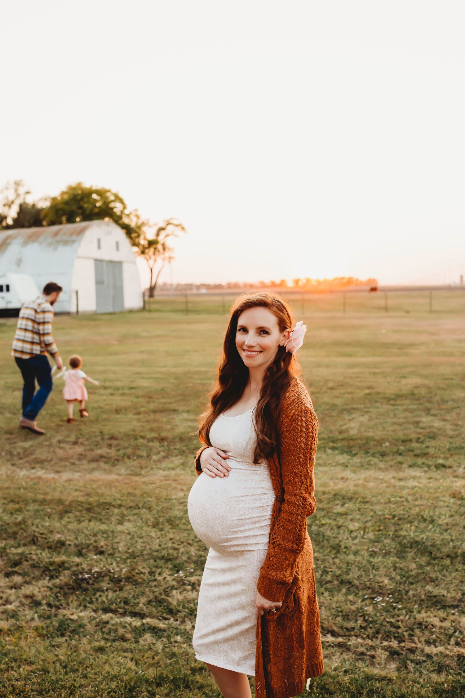 A smiling pregnant woman in a white dress and rust cardigan poses on a farm at sunset with family behind her.