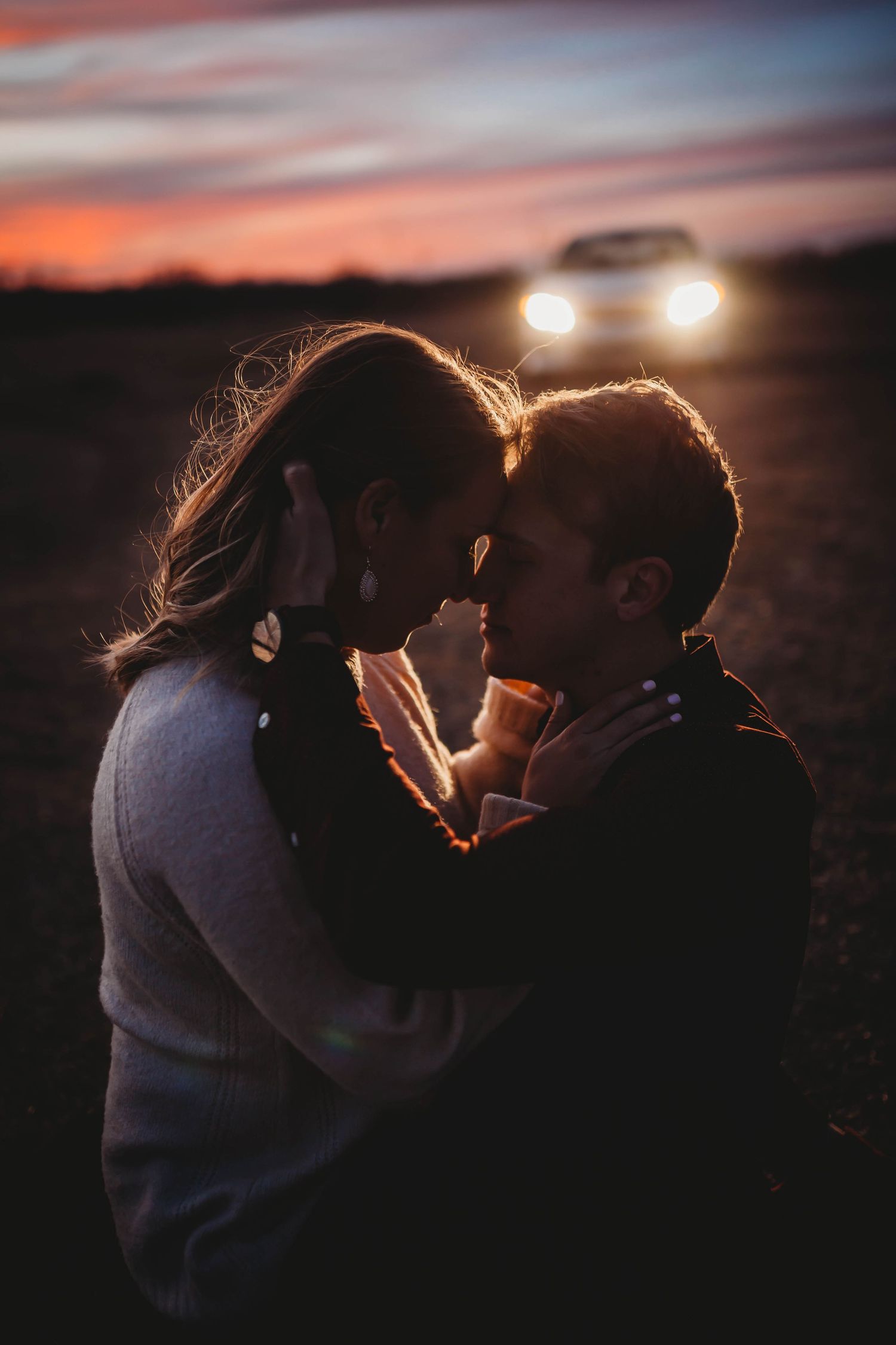 Romantic couple sharing an intimate moment at sunset with car headlights glowing in the background.