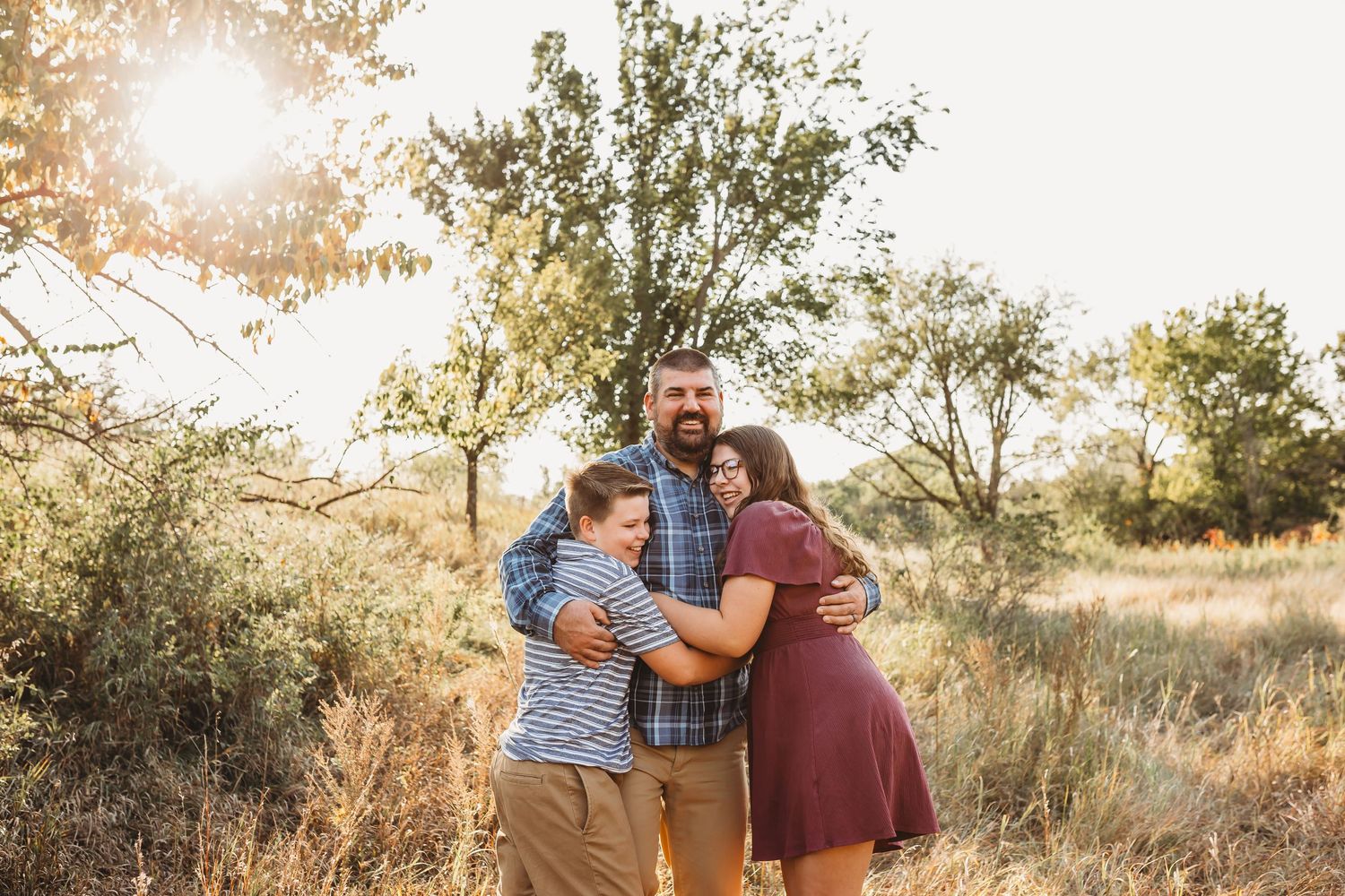 Father hugging two children in a golden sunlit field with trees in the background.