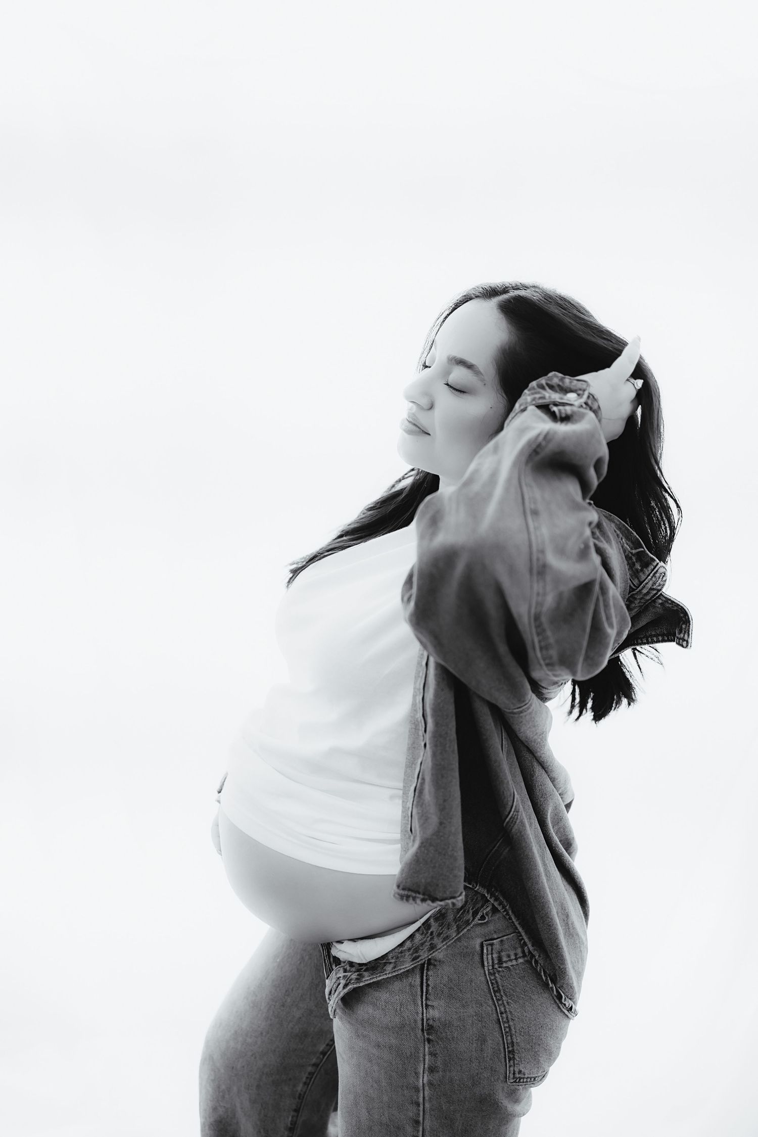 Black and white portrait of a pregnant woman in a denim jacket and jeans touching her hair outdoors.