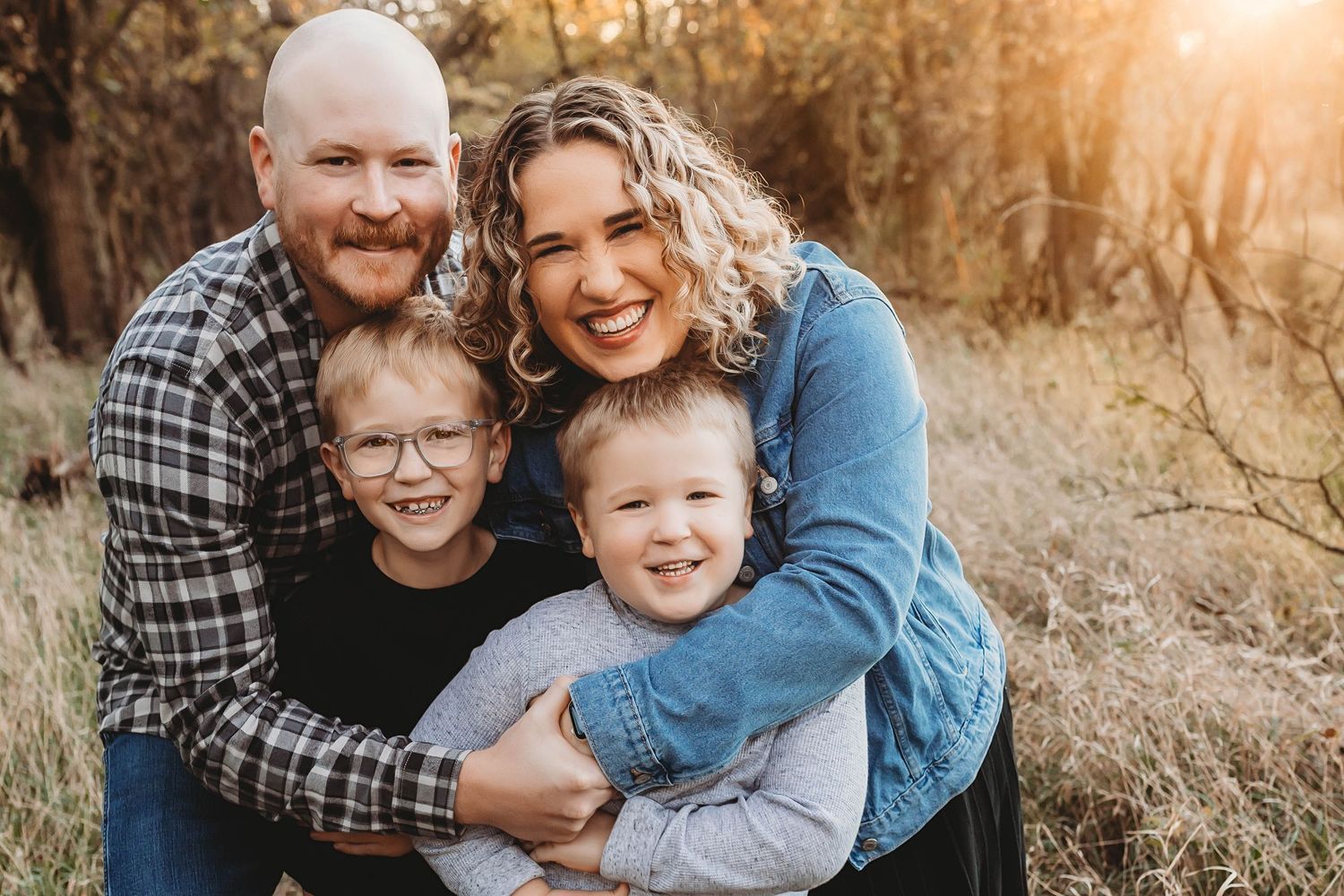 Happy family of four smiling outdoors in autumn, parents hugging two young boys in a golden field.