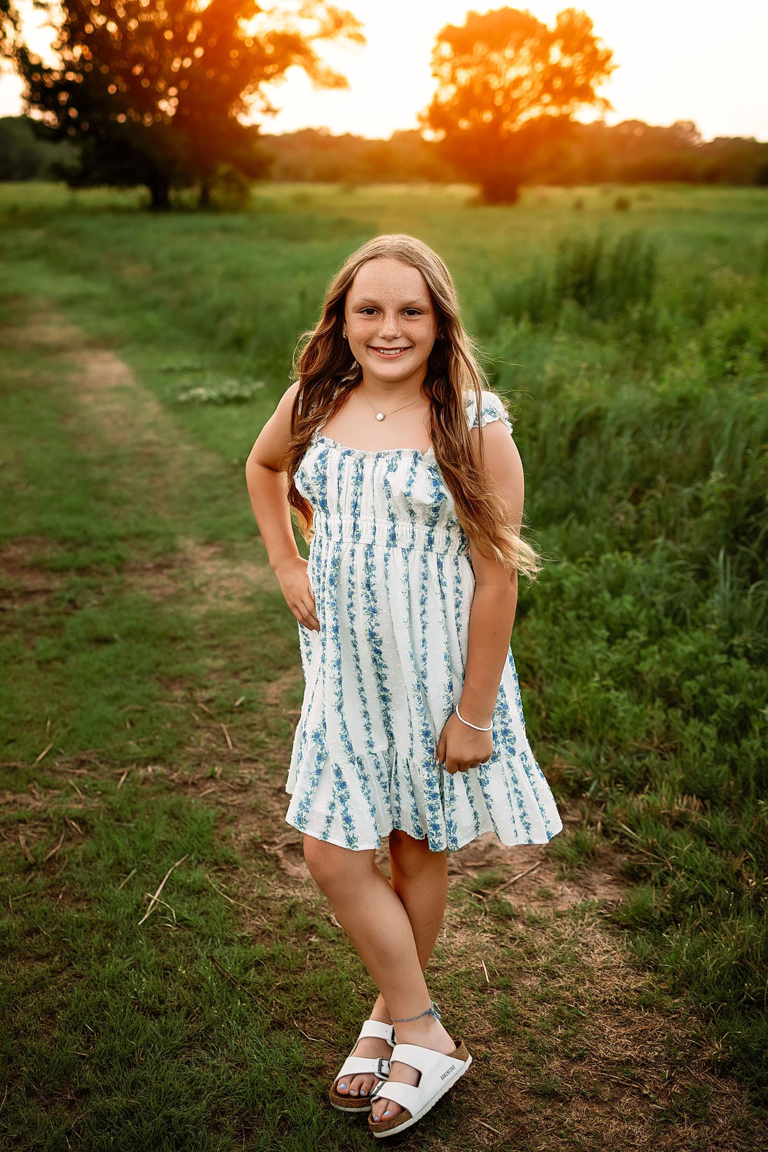 Young girl in floral sundress and white sandals smiling outdoors at golden sunset in green field.