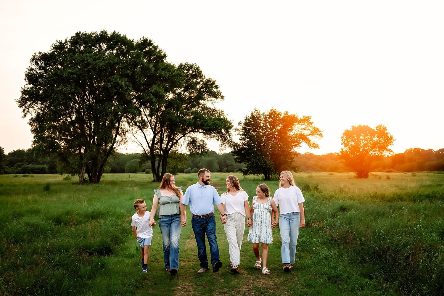 Family of six walking through a sunlit green field at golden hour with large trees in the background.