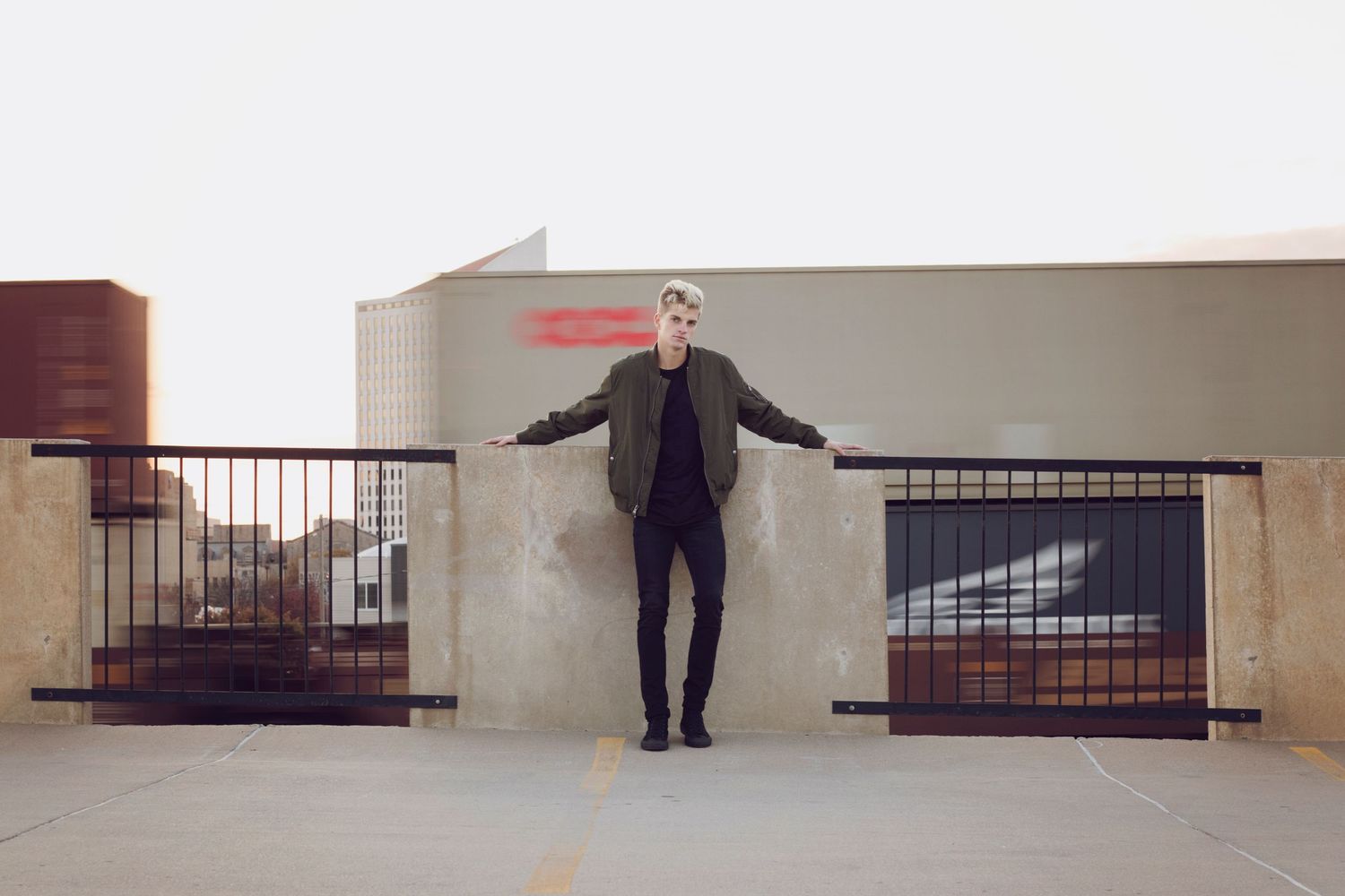 A stylish person in dark clothing and sunglasses stands on a rooftop parking structure with metal railings.