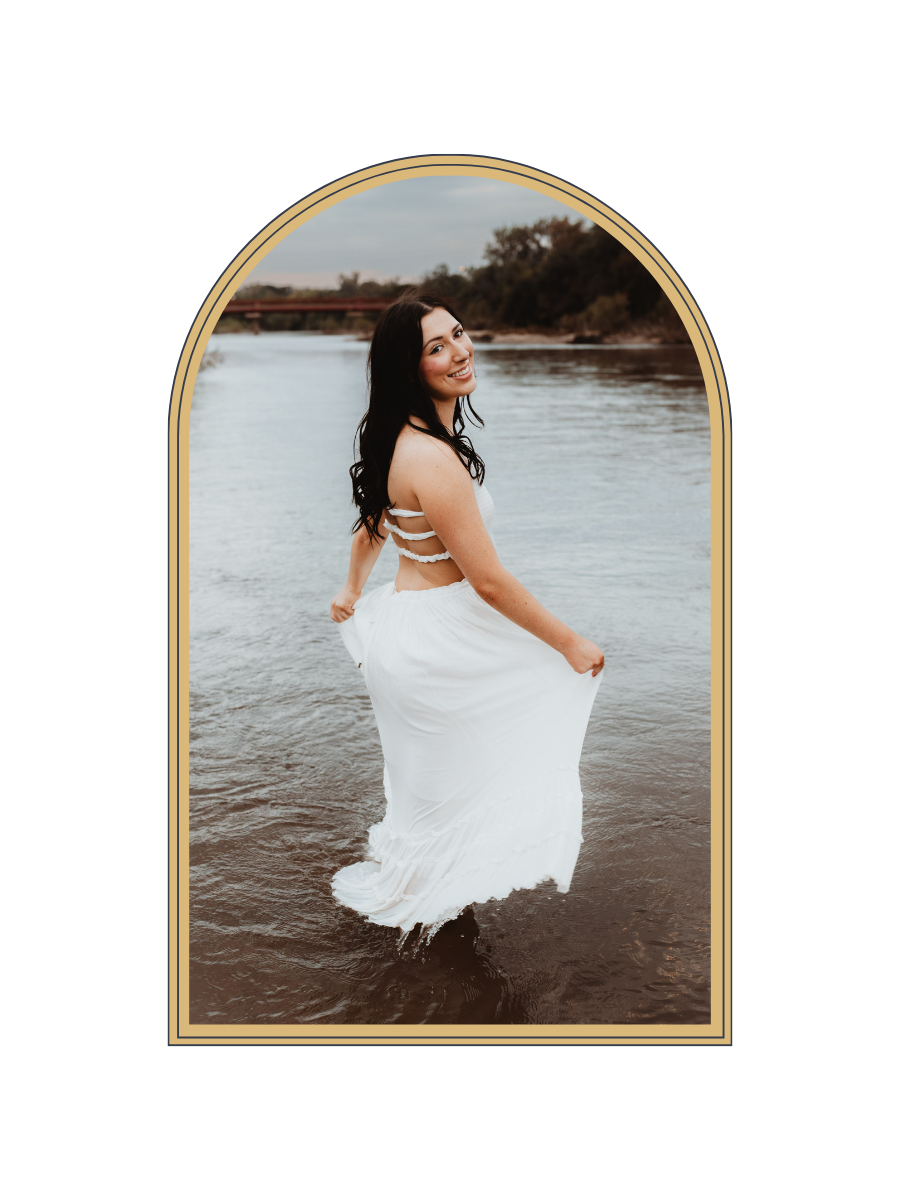 A woman in a white dress stands in shallow water, smiling over her shoulder in a serene outdoor setting.