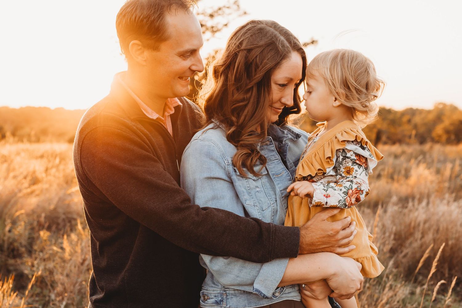 Young couple holding their toddler daughter in a golden sunlit field during fall season.