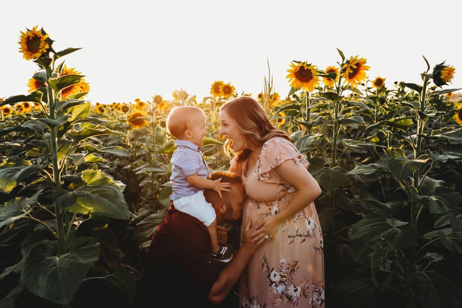 A pregnant woman in a floral dress holds a baby in a sunflower field bathed in warm golden sunset light.