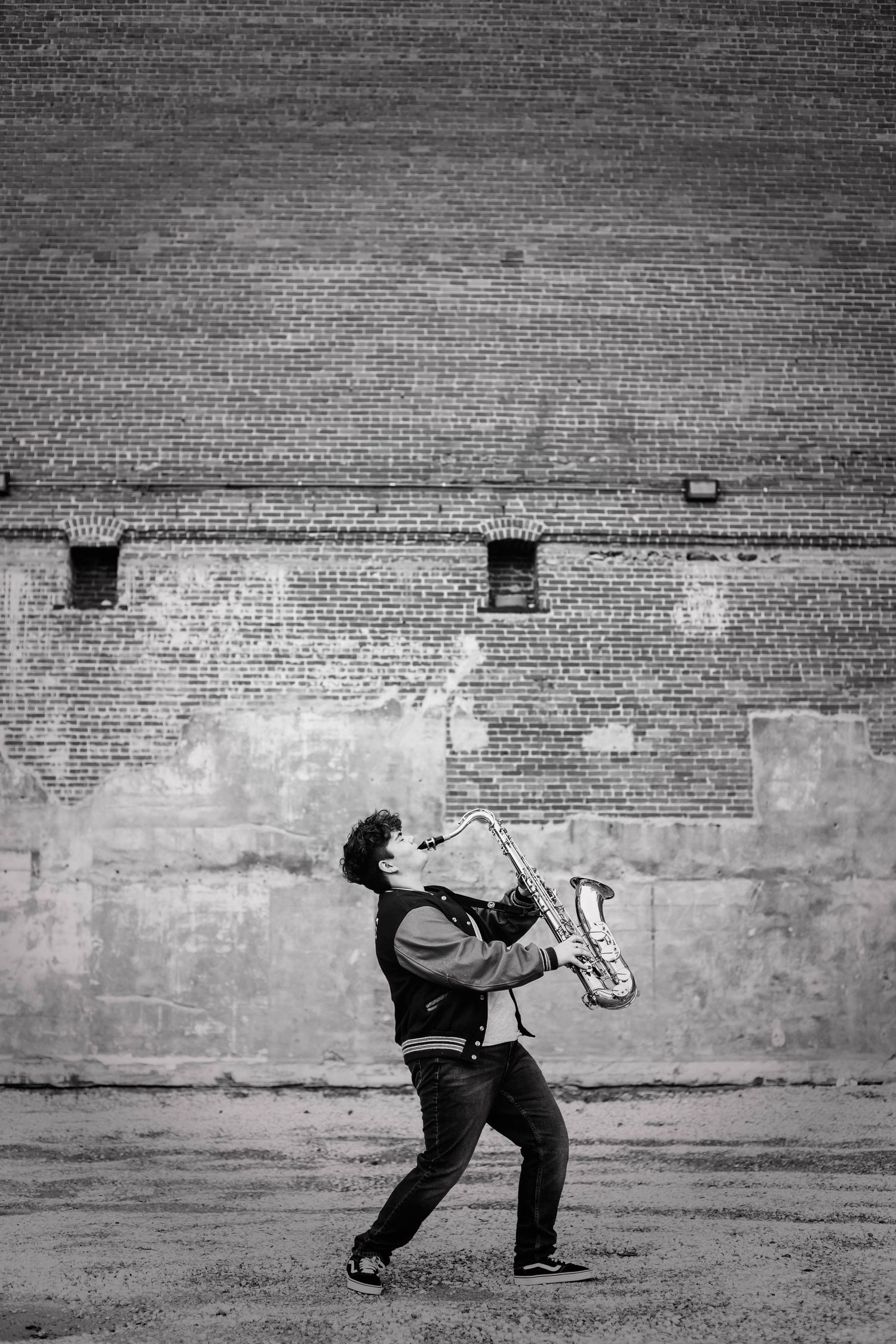 Black and white photo of musician playing saxophone outdoors against brick wall.
