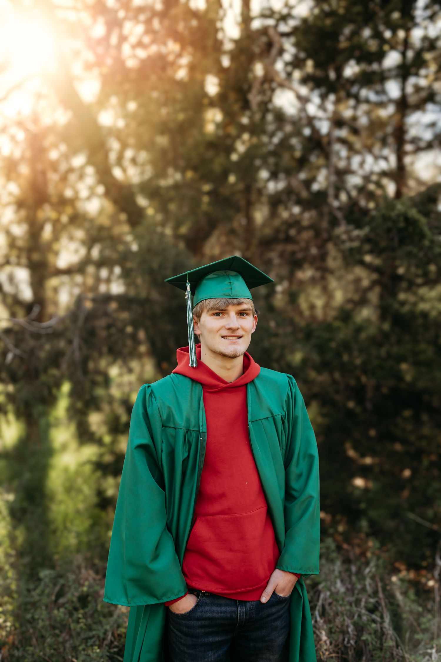 Young male graduate smiling in green cap and gown with red hoodie in sunlit forest.