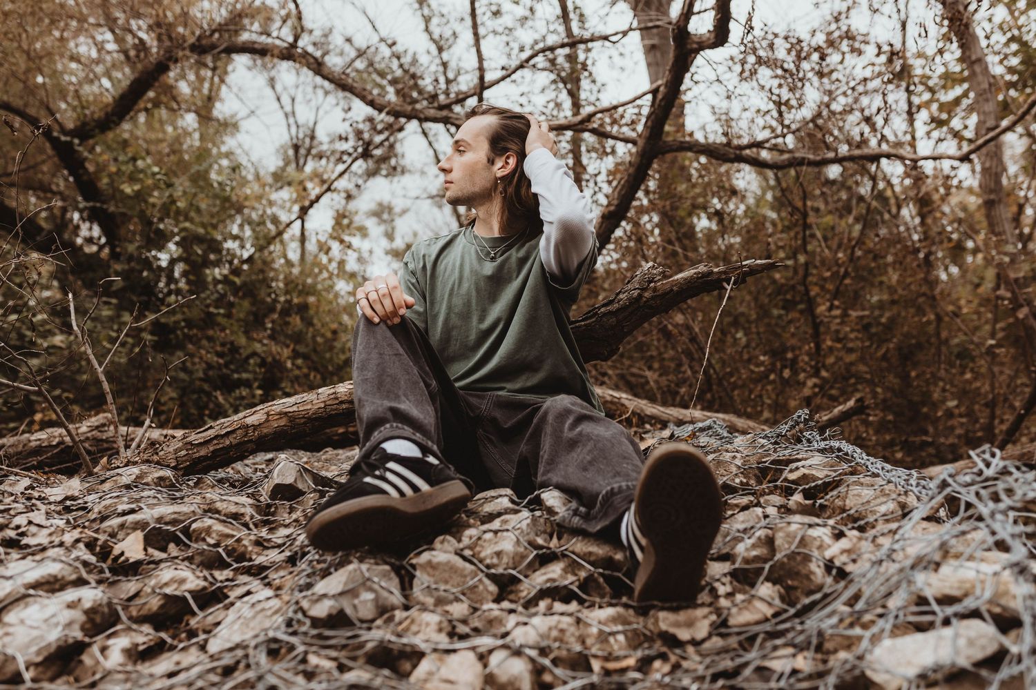 Young man sitting on rocks among bare tree branches in an autumn forest setting.