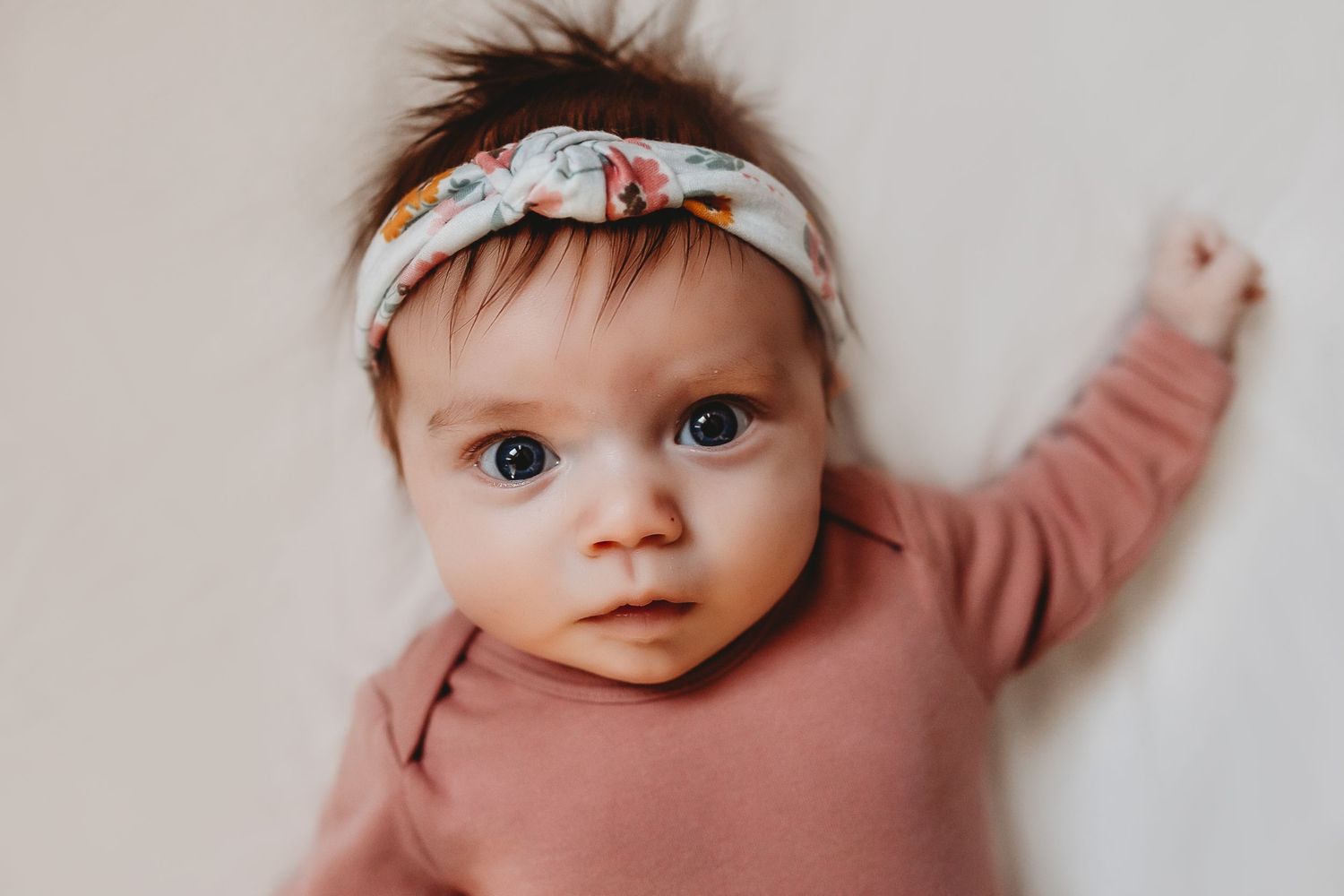 Close-up of a baby girl wearing a floral headband and pink outfit lying on a white blanket.