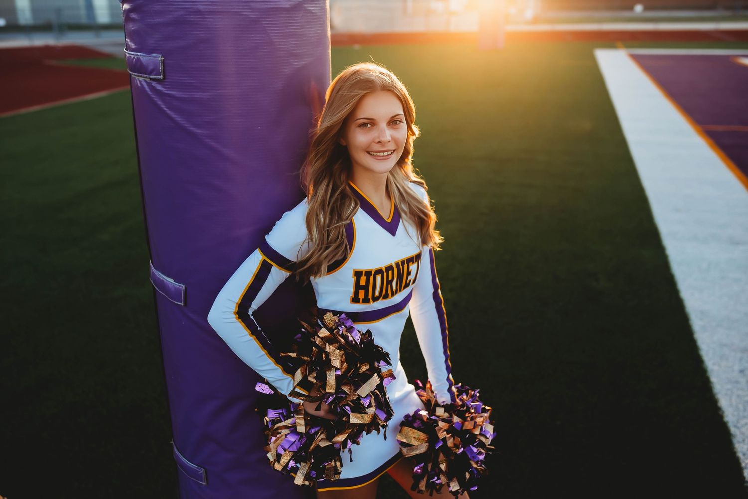 A cheerleader in a Hornets uniform smiles on a football field at sunset holding purple and gold pom-poms.