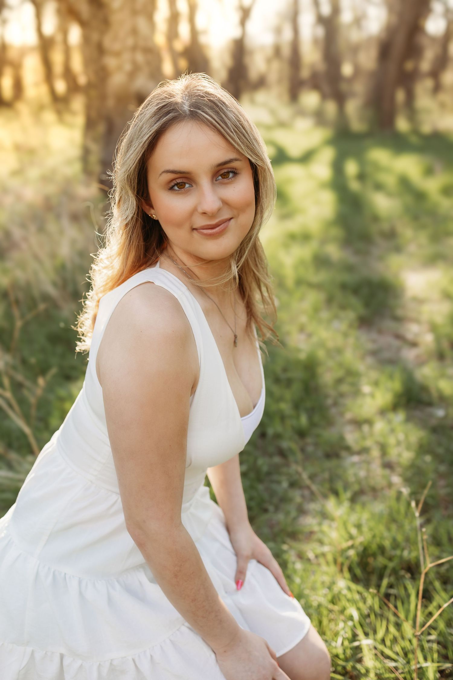 A smiling blonde woman in a white sundress sits outdoors in a sunlit forest during golden hour.