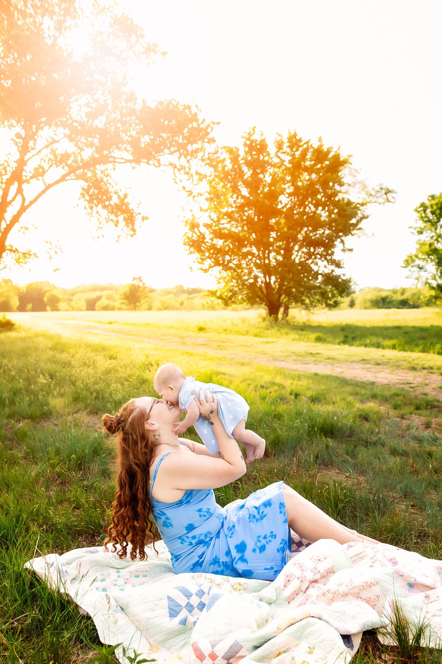 Mother in blue floral dress lifting baby outdoors on a quilt in a sunlit green field with trees at golden hour.