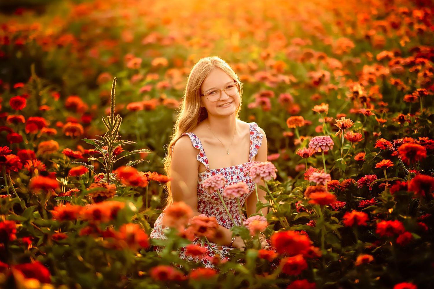 Young blonde woman smiling in a vibrant red flower field, wearing a floral dress, bathed in warm golden sunlight.
