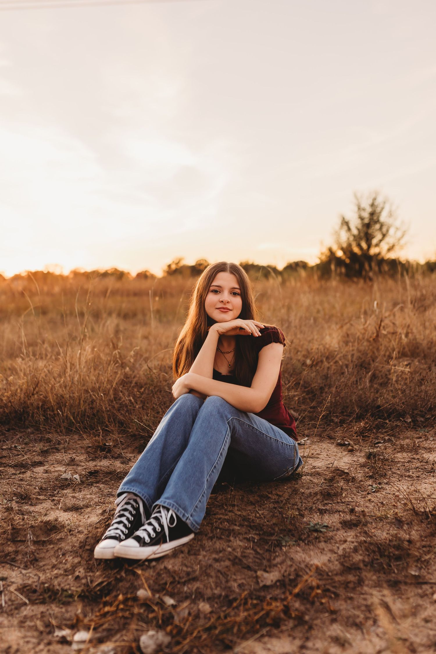 Young woman sitting in golden field at sunset, wearing jeans and Converse sneakers, smiling warmly at camera.