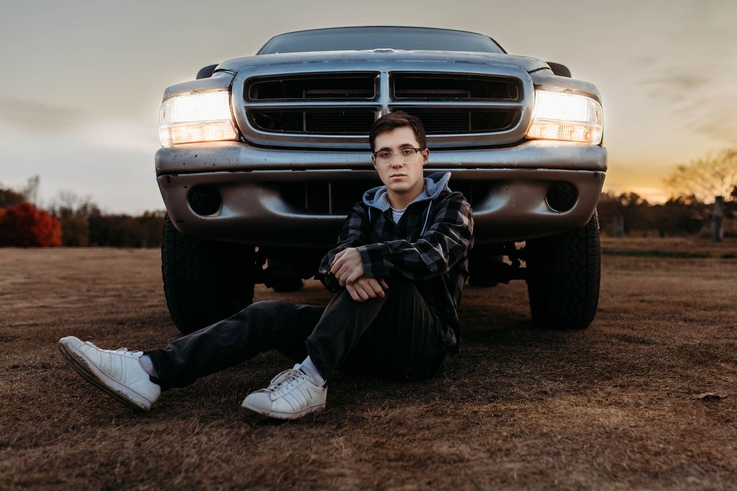 Young man in leather jacket sitting in front of a lifted truck at sunset in a rural field.