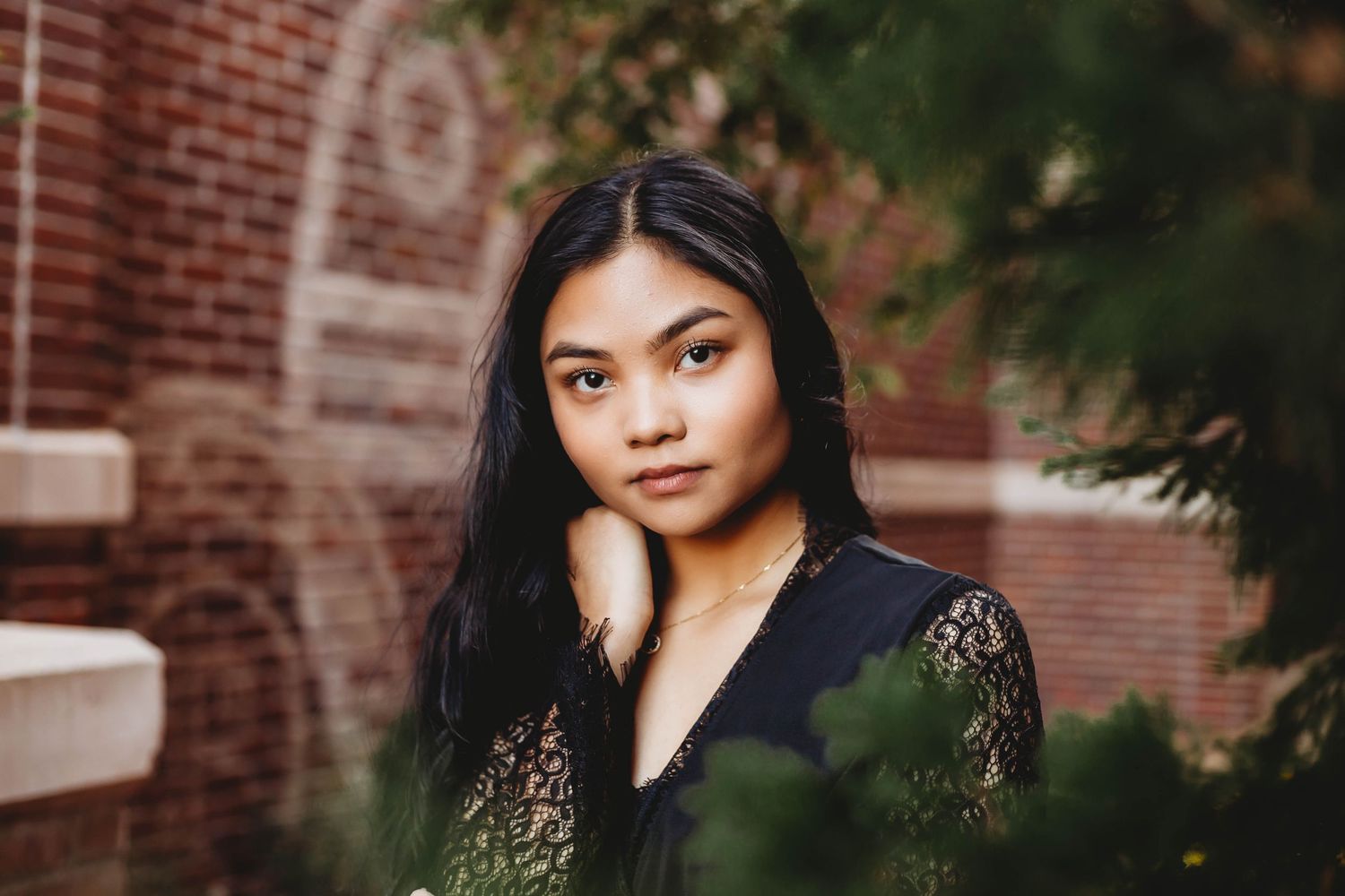 Teen girl with long dark hair posing near greenery and brick wall.
