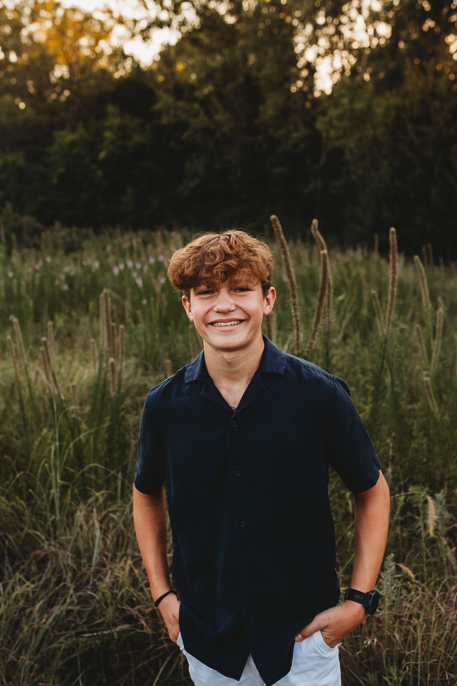 Smiling teenage boy with curly red hair posing in field at golden hour.