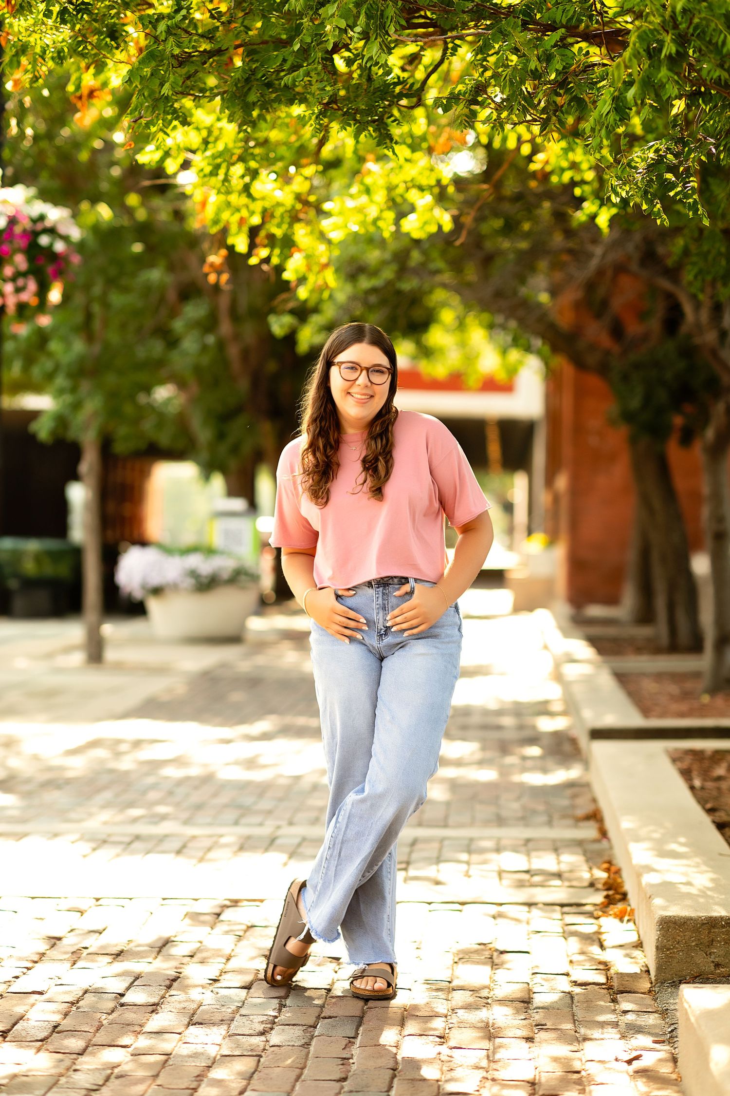 Young woman in pink top and jeans smiling outdoors on brick path under green trees.