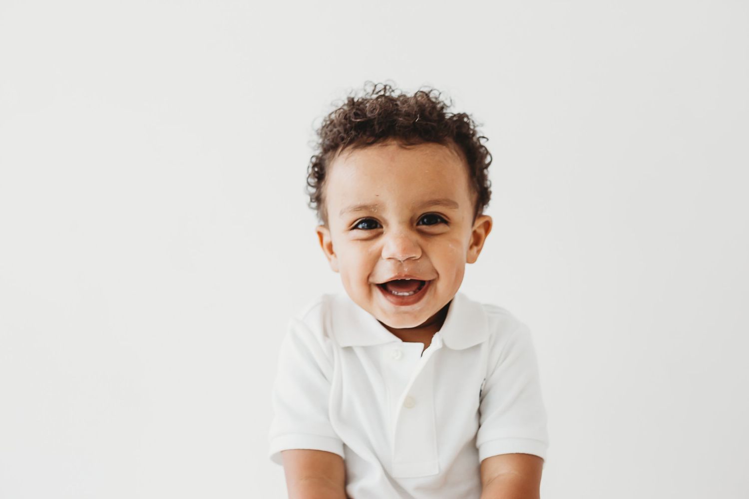 Adorable smiling baby boy with curly hair wearing a white shirt on white background.