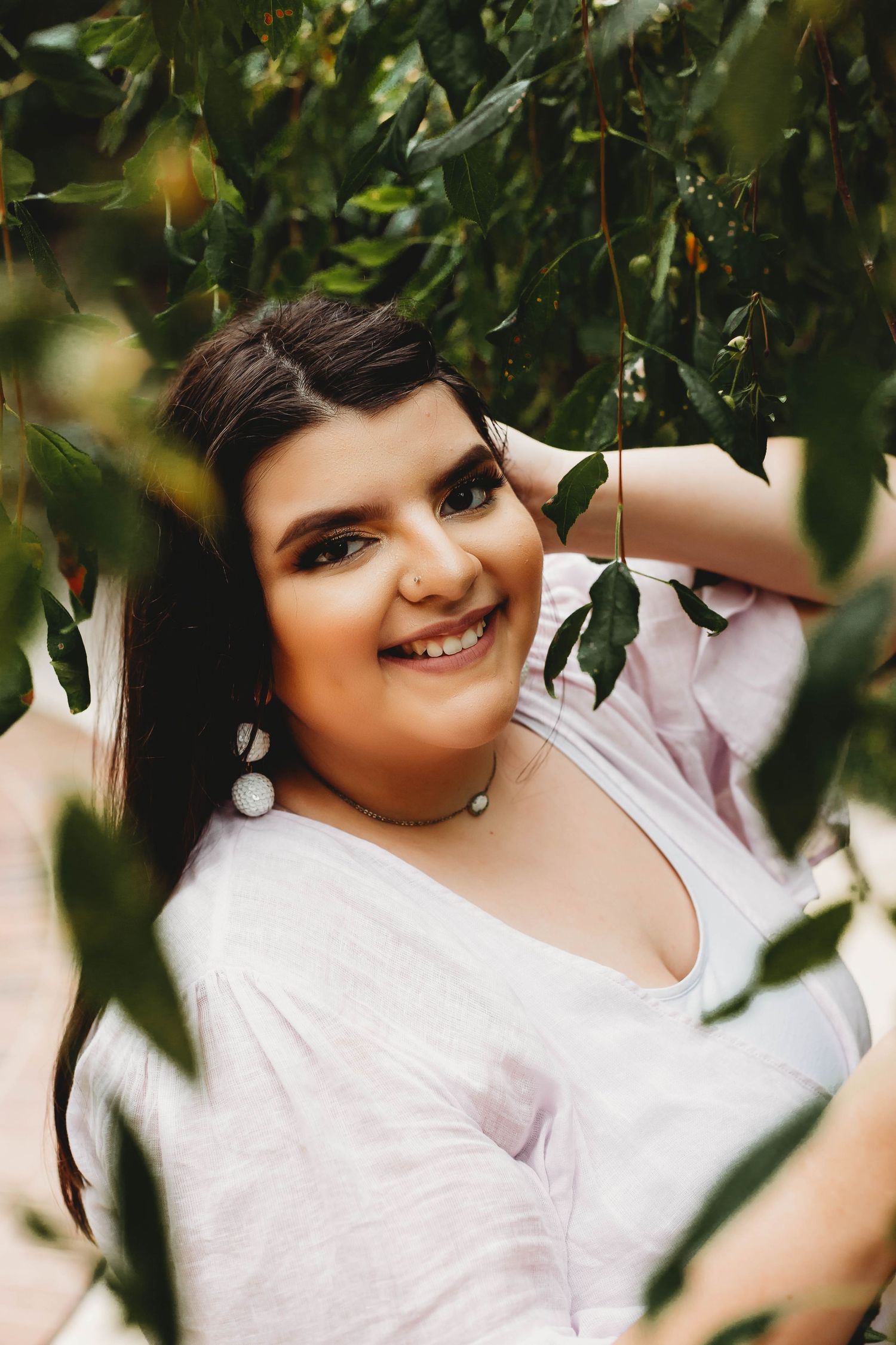 Smiling young woman with dark hair posing among lush green leaves in natural setting.