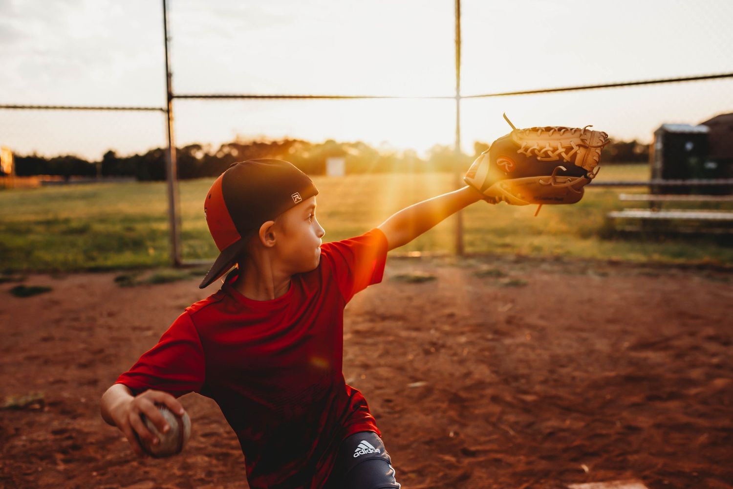 Young baseball player catching a ball at sunset on a dirt field wearing red uniform.
