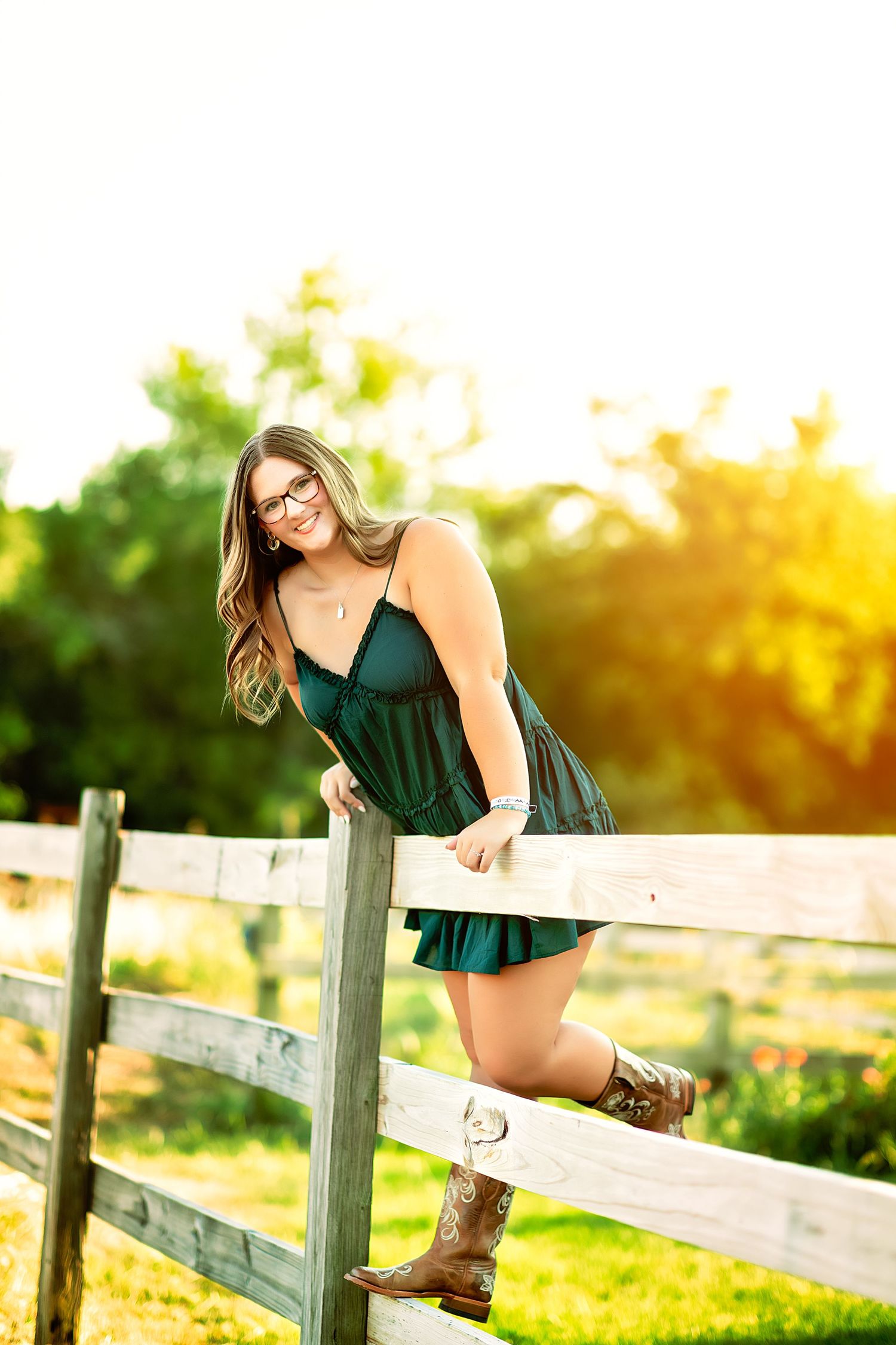 Young woman in green dress and cowboy boots leaning on a wooden fence in a sunny outdoor setting.
