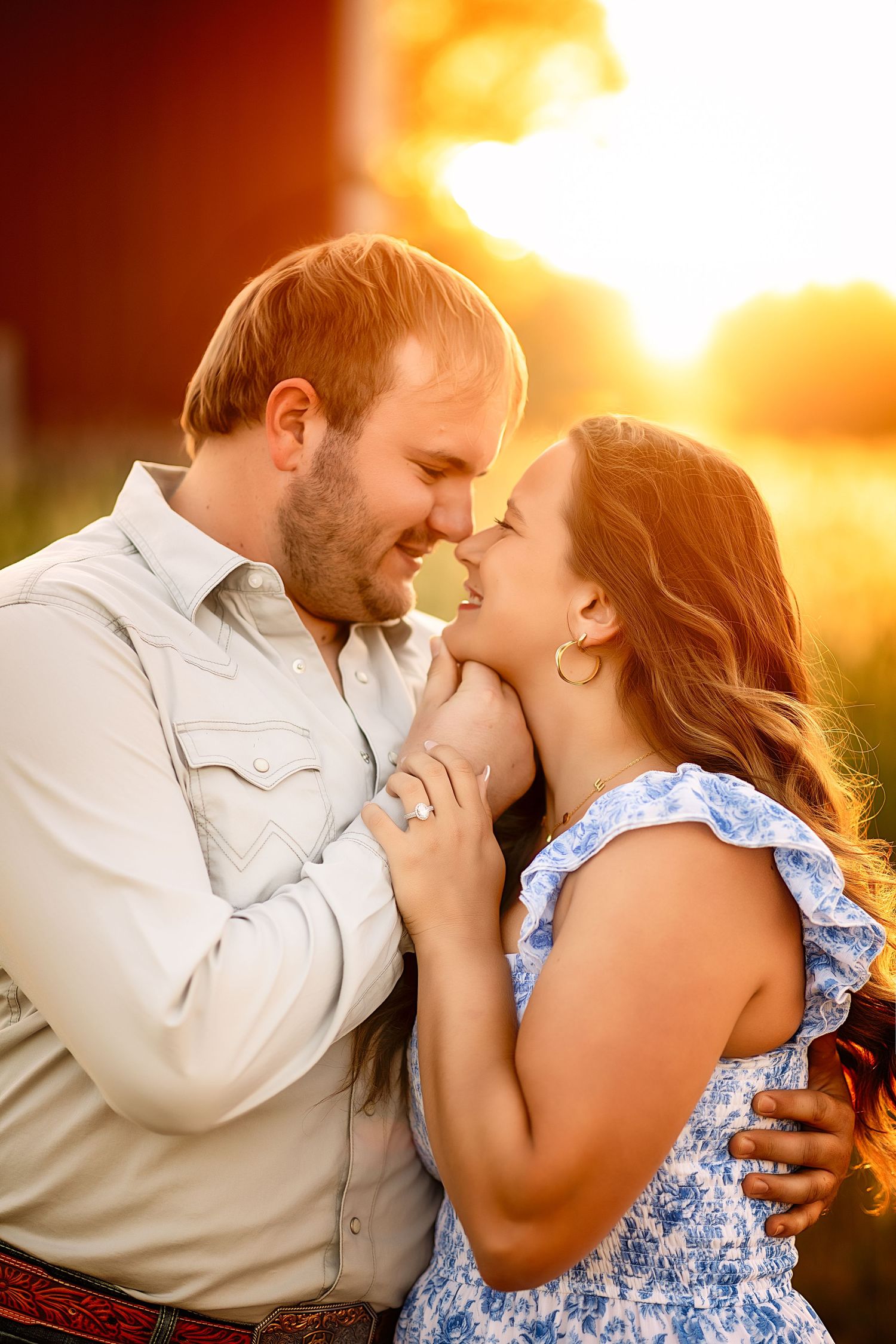 A couple shares a romantic moment at golden hour, nearly kissing outdoors near a red barn.