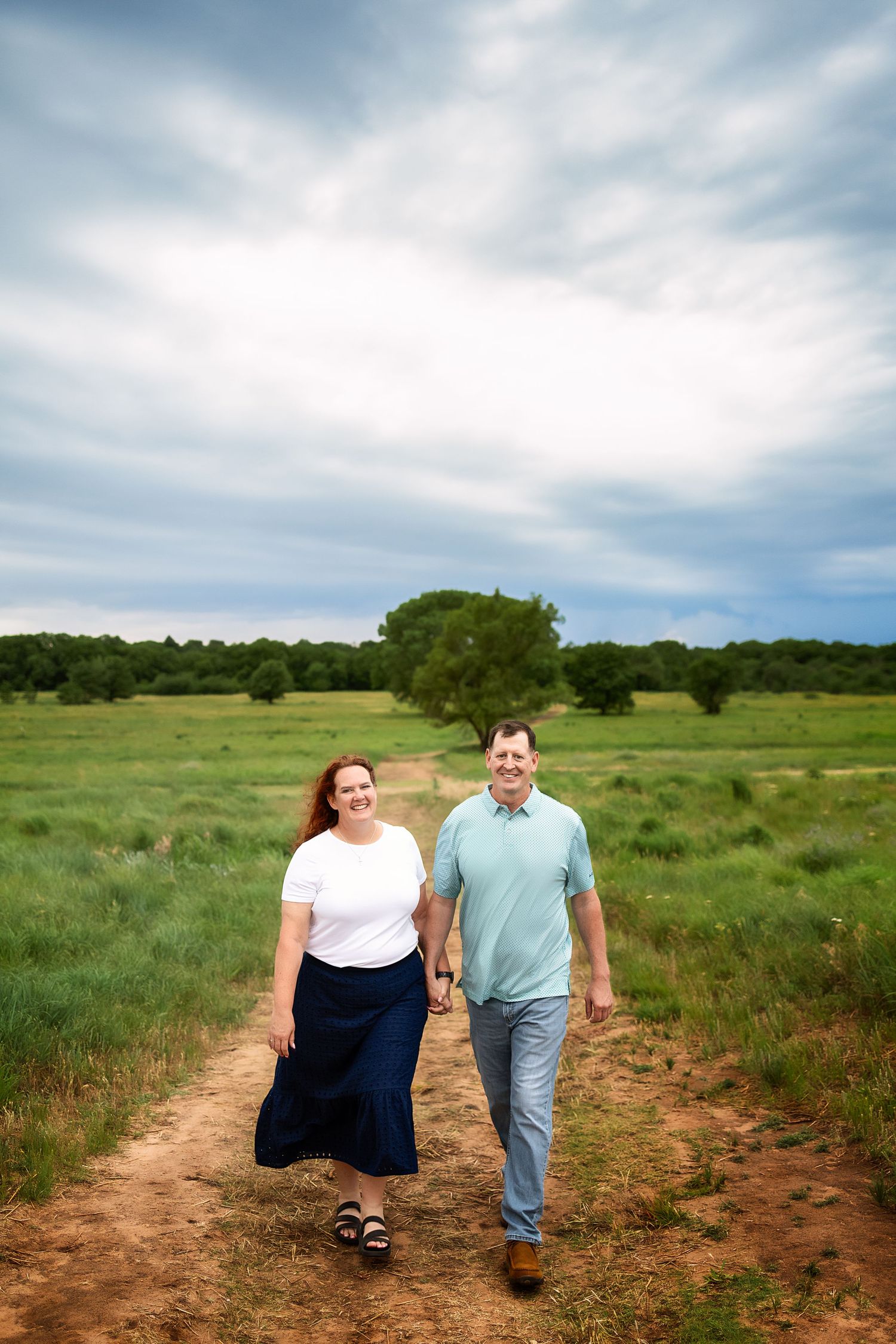 Couple holding hands walking on a dirt path through a lush green field under a dramatic cloudy sky.