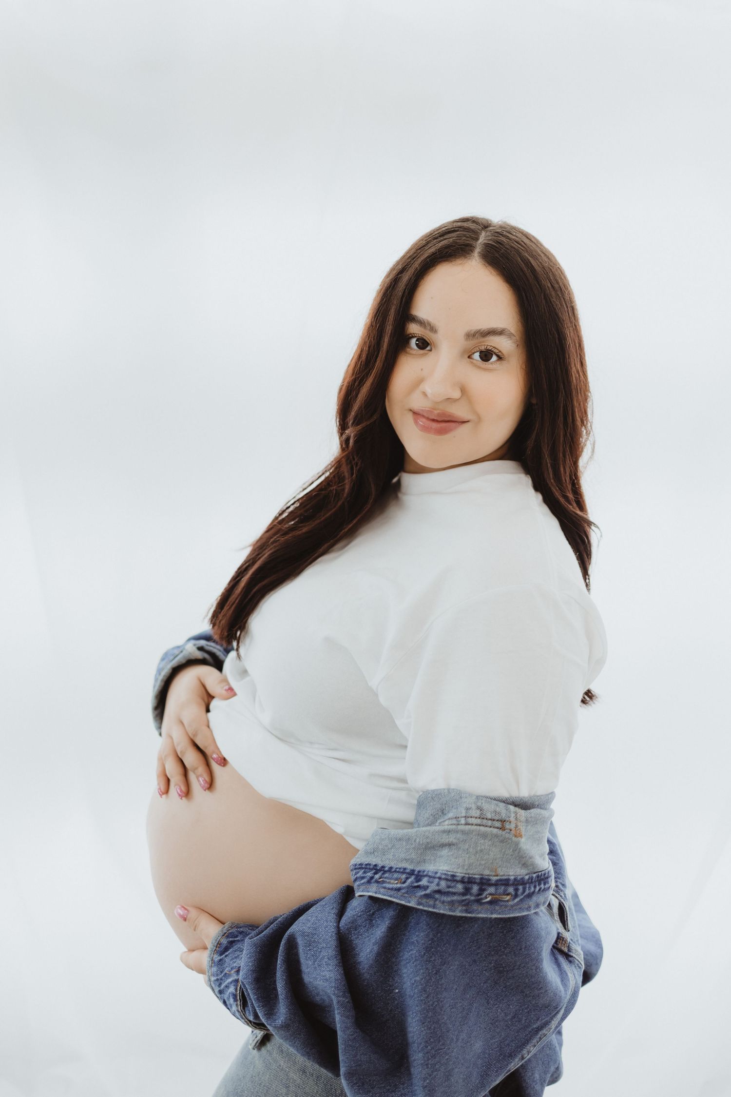 Smiling pregnant woman in white top and denim jacket holds her bare belly sideways against white backdrop.
