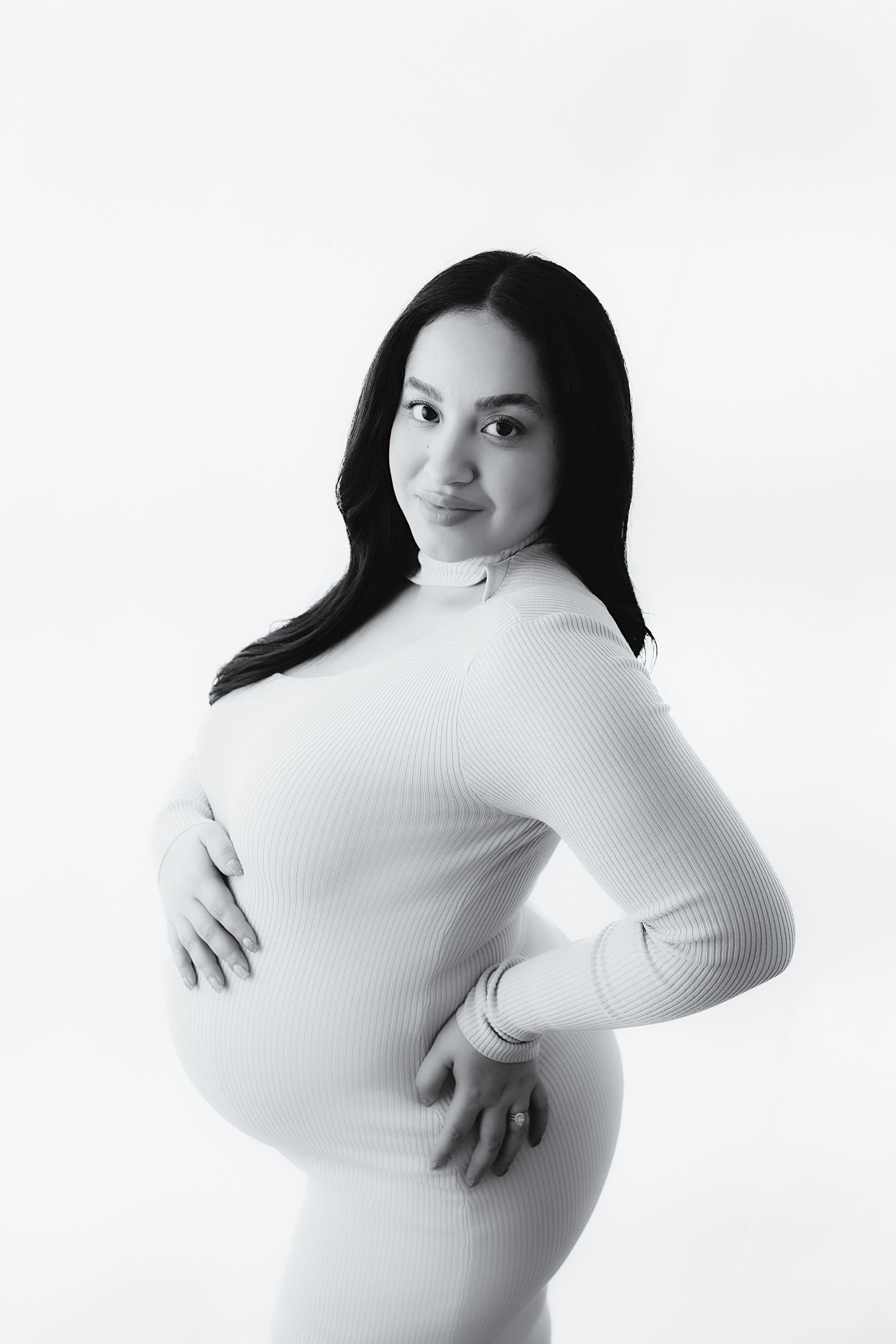 Black and white portrait of smiling pregnant woman in white dress holding baby bump.