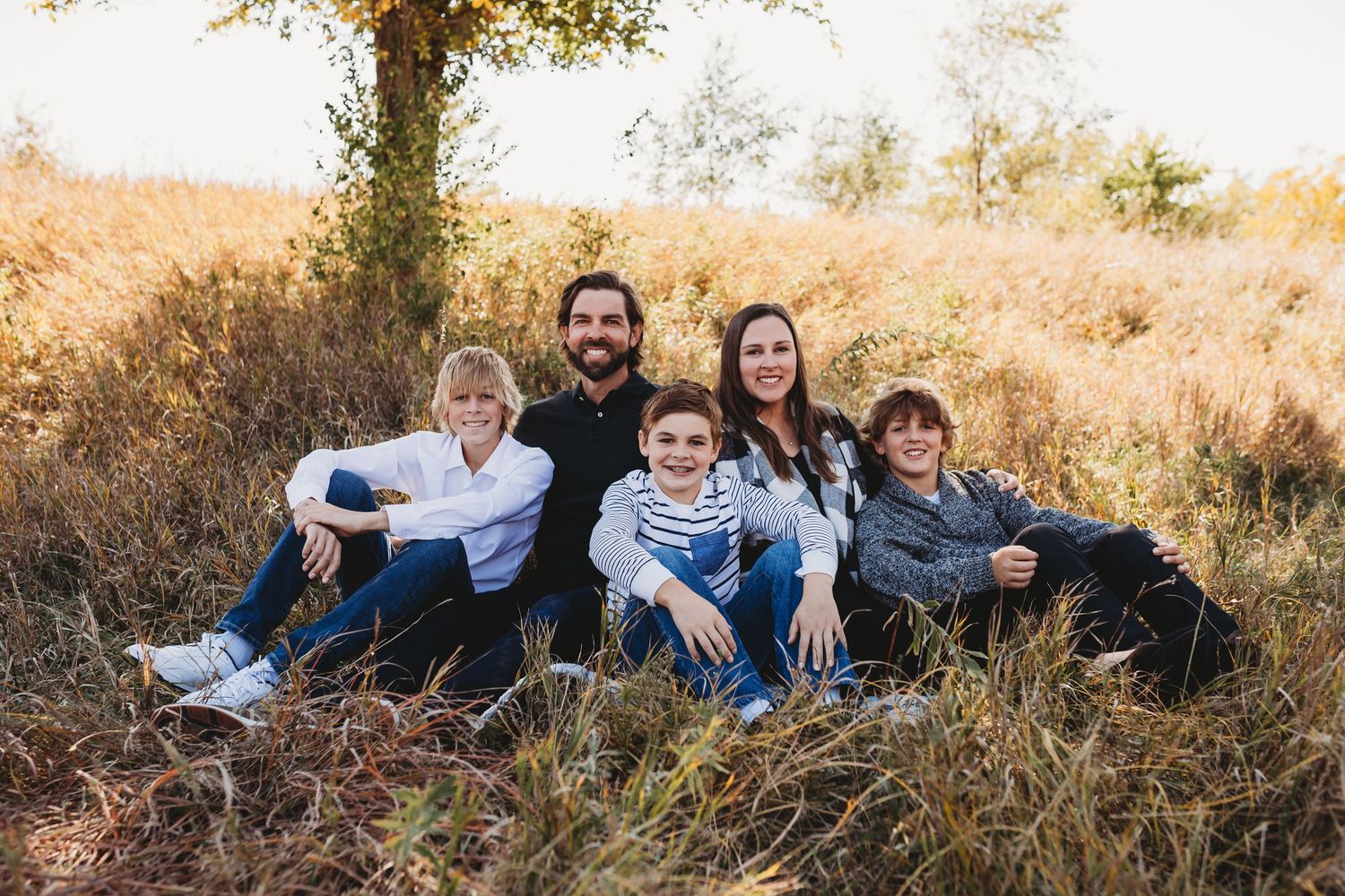Family of five relaxing together in golden autumn field, smiling warmly at the camera.