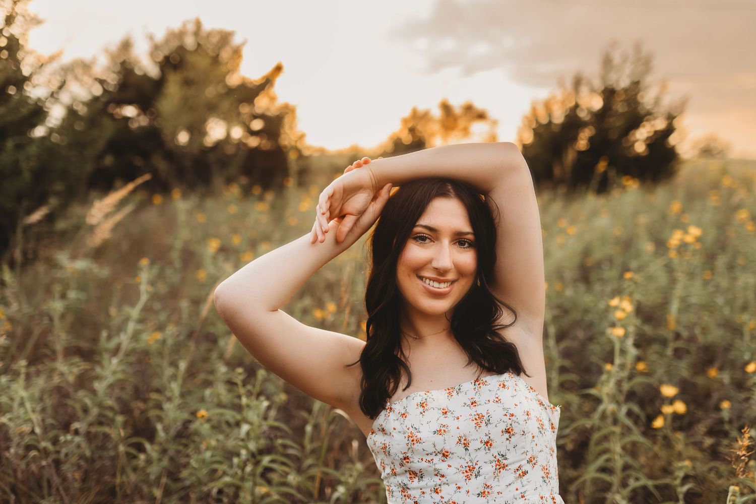 Young woman in floral dress smiling in golden hour sunflower field with arms raised.
