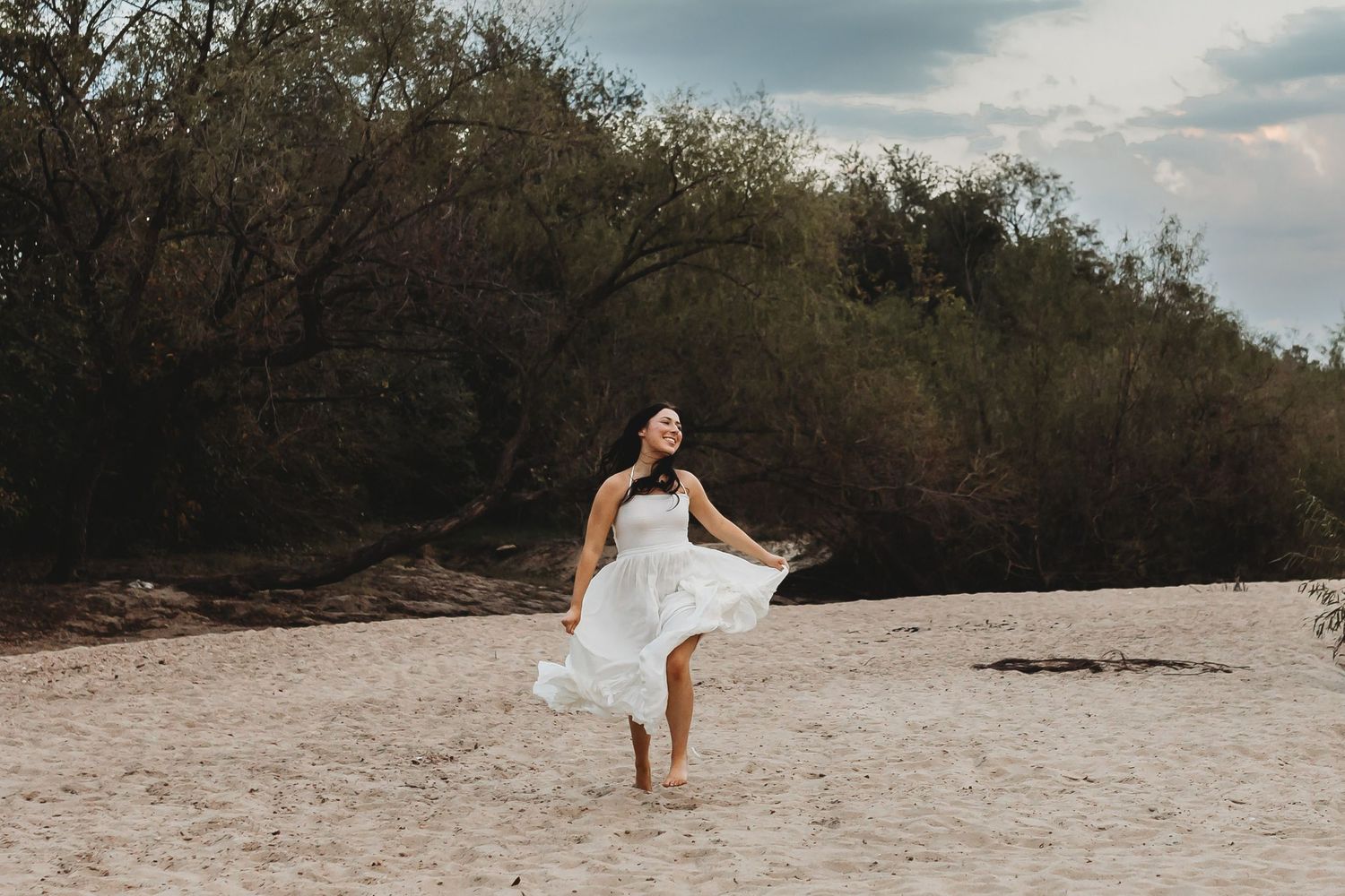 A woman in a flowing white dress dances barefoot on sandy beach with lush green trees in the background.