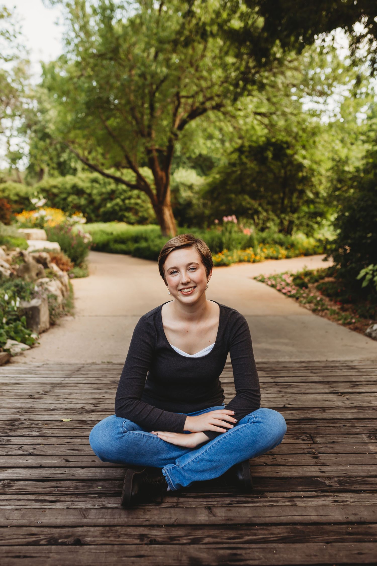A smiling young woman with short hair sits cross-legged on a wooden boardwalk in a lush green garden park.