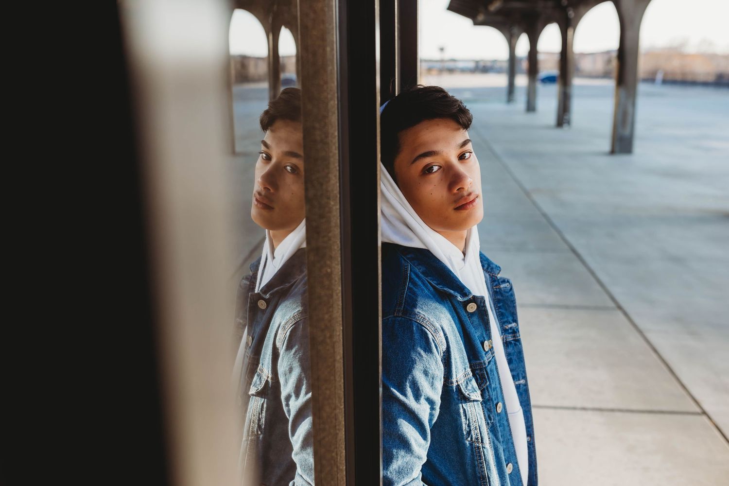 Young man in denim jacket leaning against reflective glass pillar in covered outdoor space.