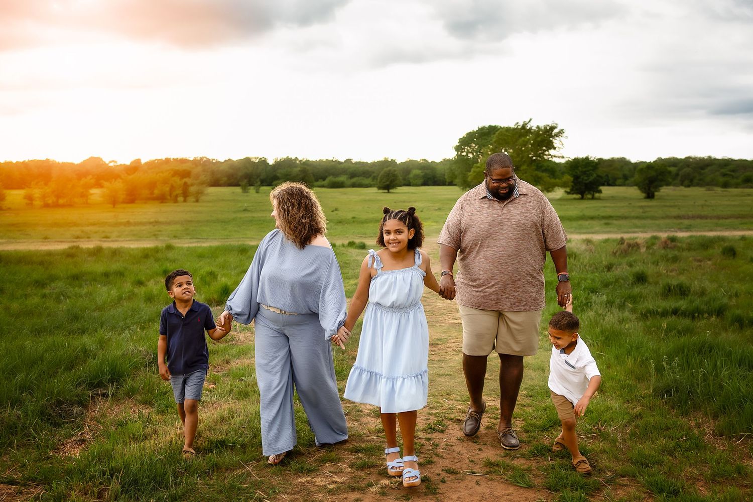 Family of five holding hands walking through a green field at sunset in a heartwarming outdoor portrait.