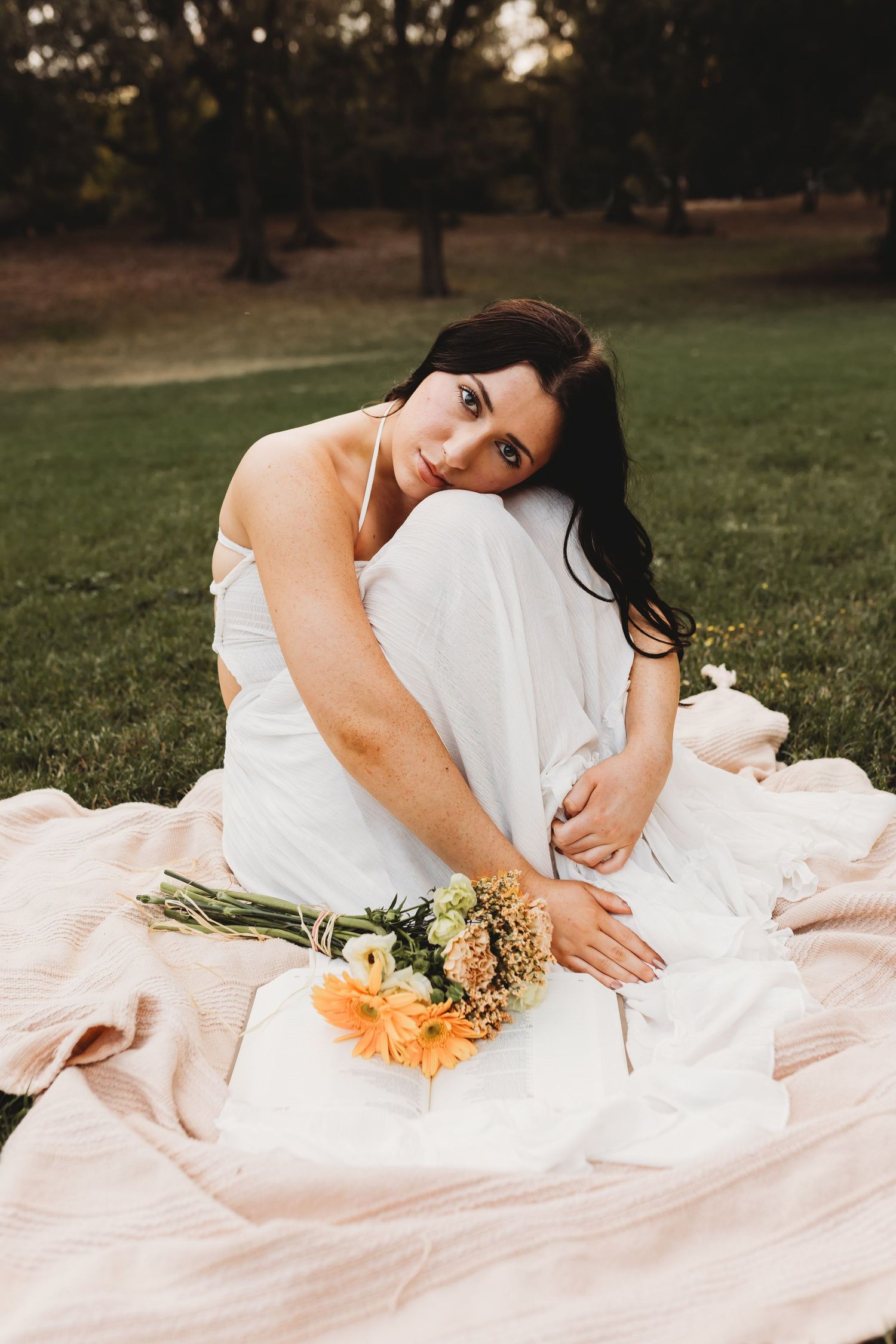 Woman in white dress sitting on pink blanket in park, holding orange and yellow flowers, looking at camera.