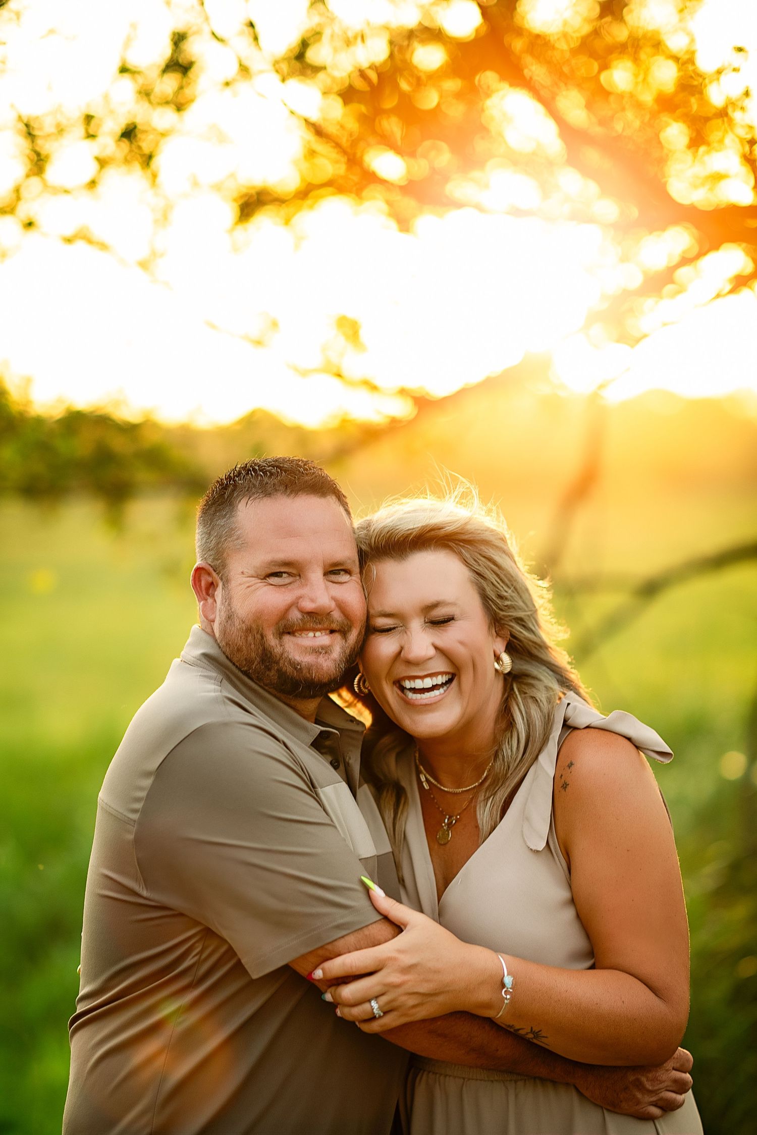 A joyful couple embraces outdoors at golden hour, laughing together in a sunlit green field.