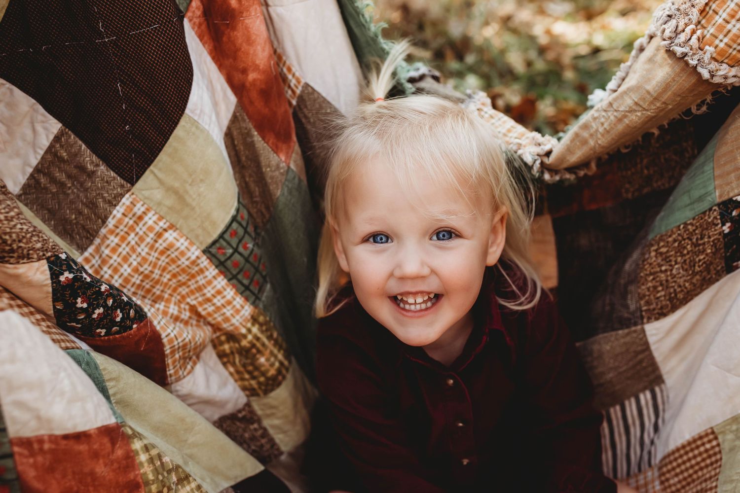 Happy blonde toddler peeking out from beneath a colorful patchwork quilt outdoors in autumn.