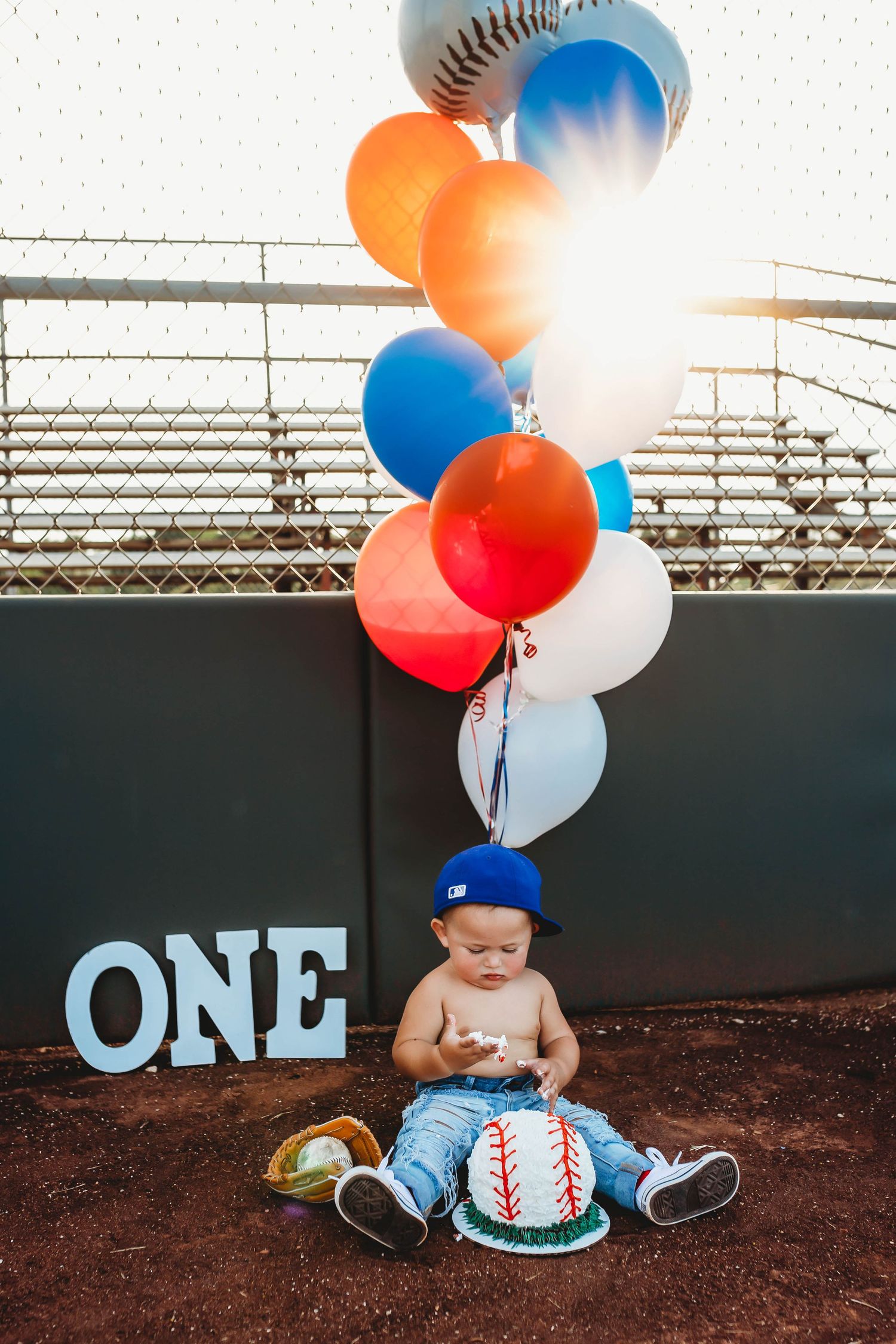 Baby boy in blue cap sits with baseball cake and colorful balloons beside ONE sign at baseball field first birthday.